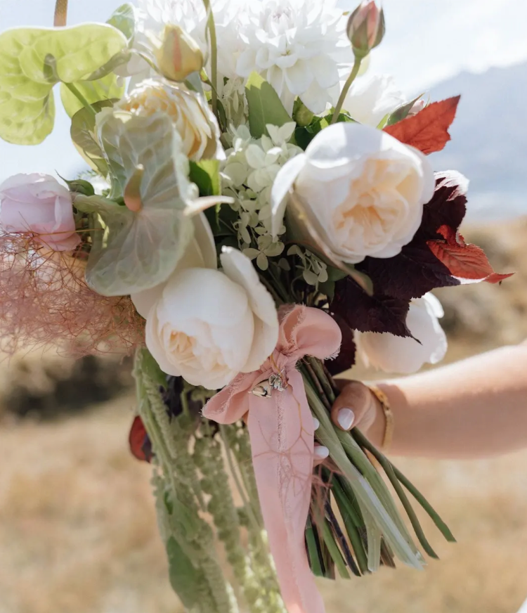 Person holding a bouquet of white and pink roses, greenery, and autumn leaves, tied with a pink ribbon.