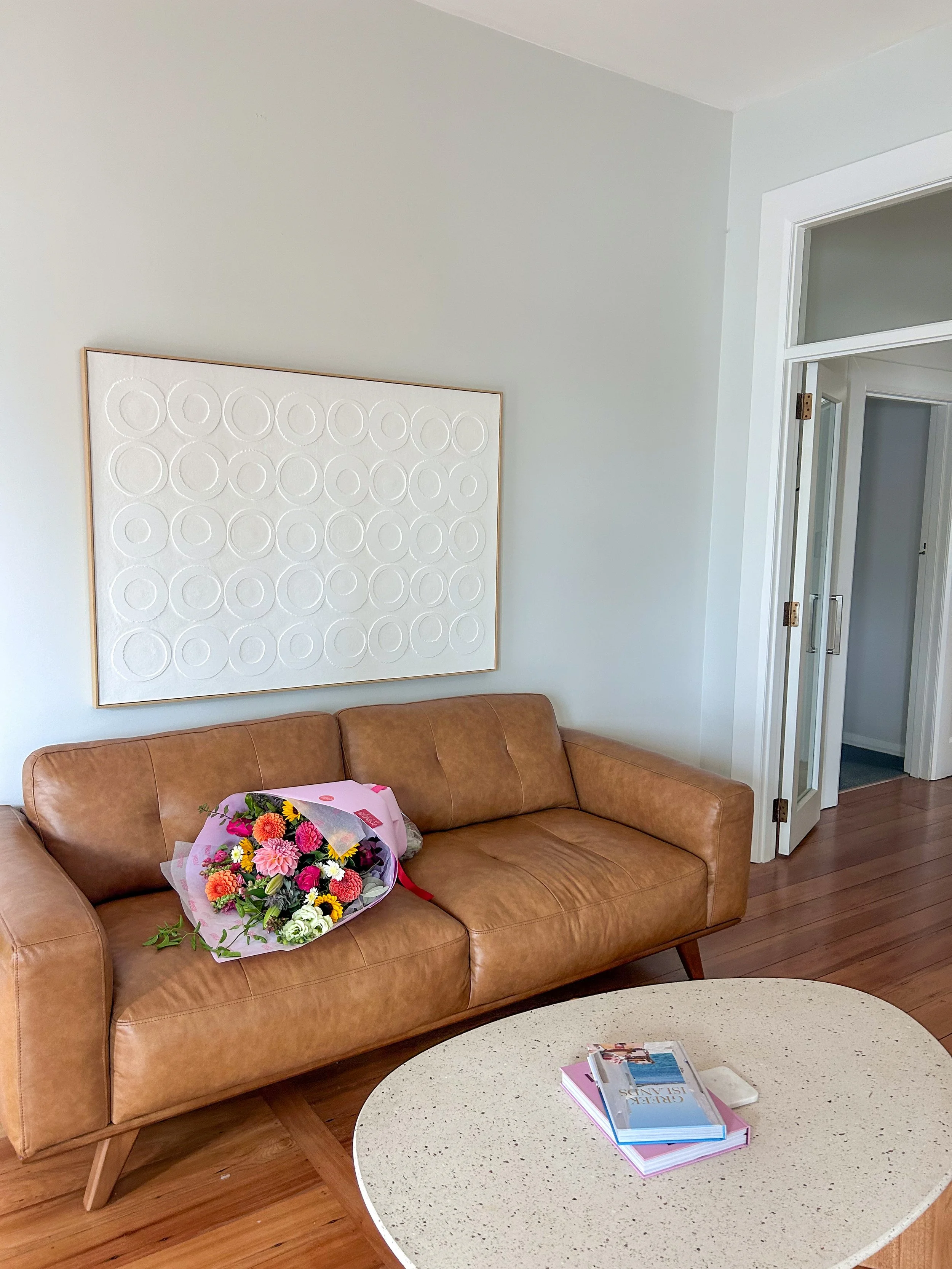 Living room with a brown leather sofa, a bouquet of colorful flowers, a white oval coffee table with books and a magazine, and a white textured wall art. White walls and hardwood floors.