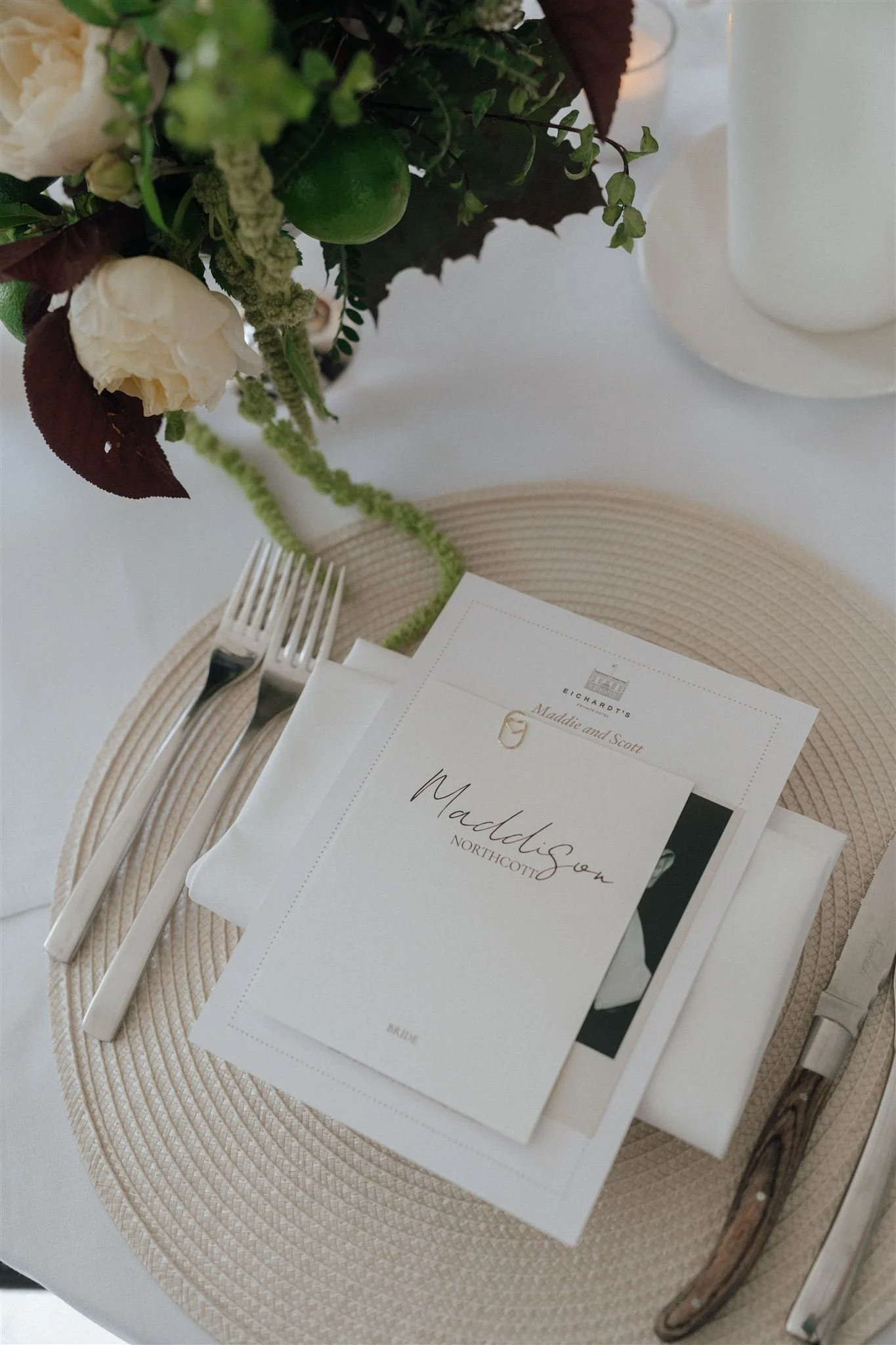 A place setting with a white plate, beige woven charger, silver cutlery, a white napkin, and a floral centerpiece with white flowers, green and dark purple leaves, and small green citrus fruits. There are also printed materials, including an invitati