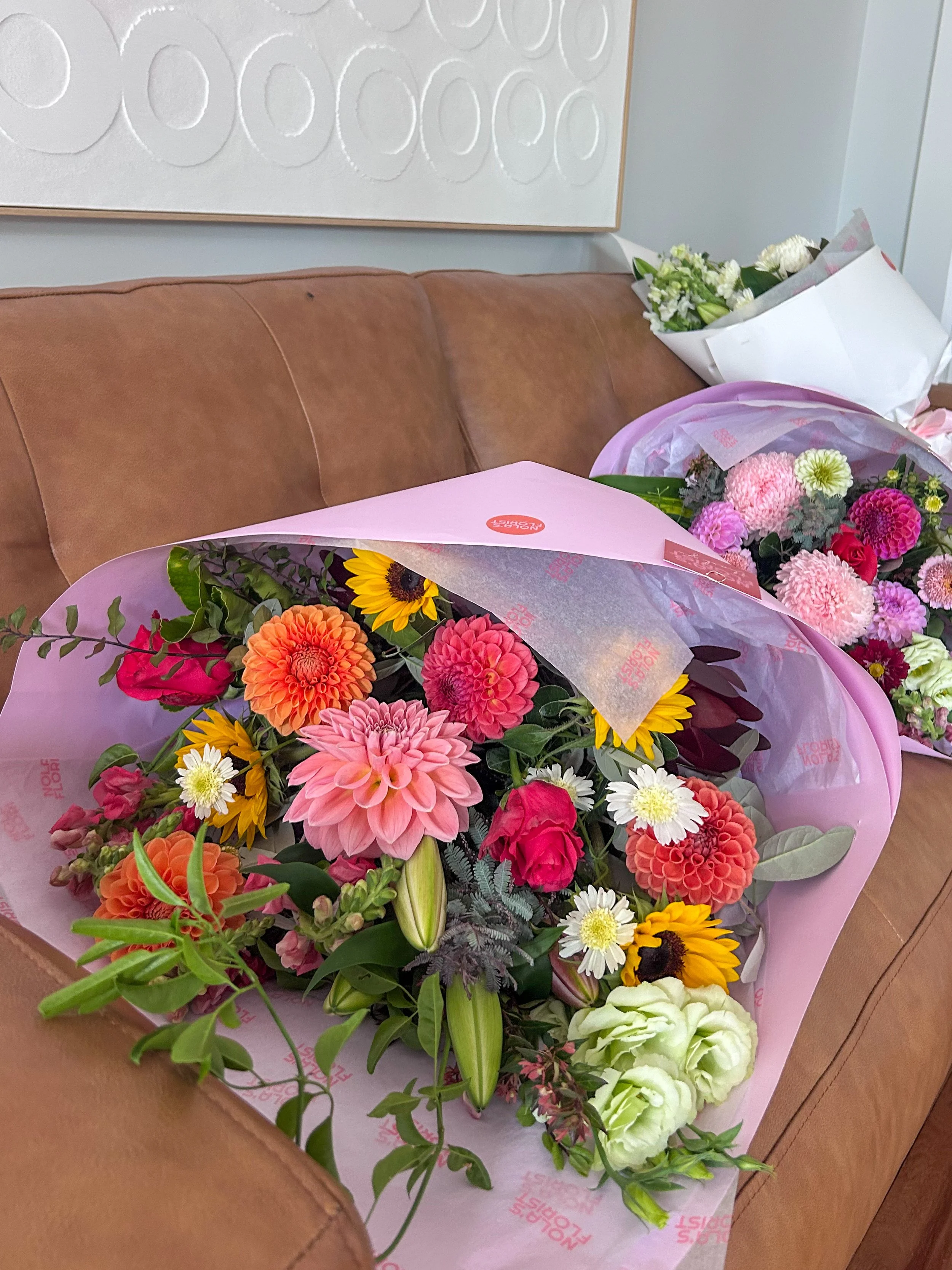 Colorful bouquet of flowers, including dahlias, sunflowers, roses, daisies, and lilies, wrapped in pink paper and placed on a brown leather couch.