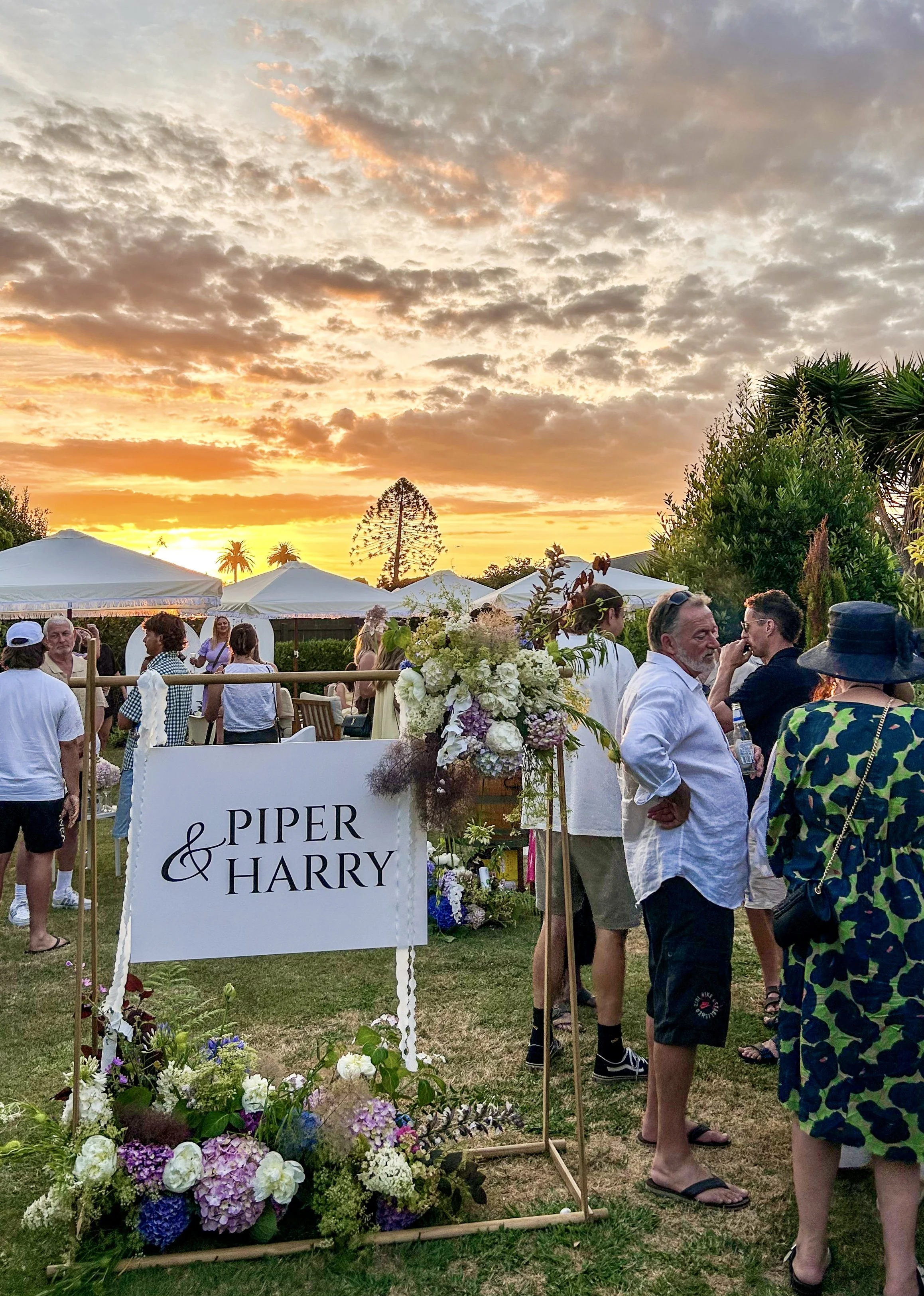 People gathered outdoors during sunset at a wedding or event, with a sign reading 'Piper & Harry' adorned with flowers, tents, and trees in the background.