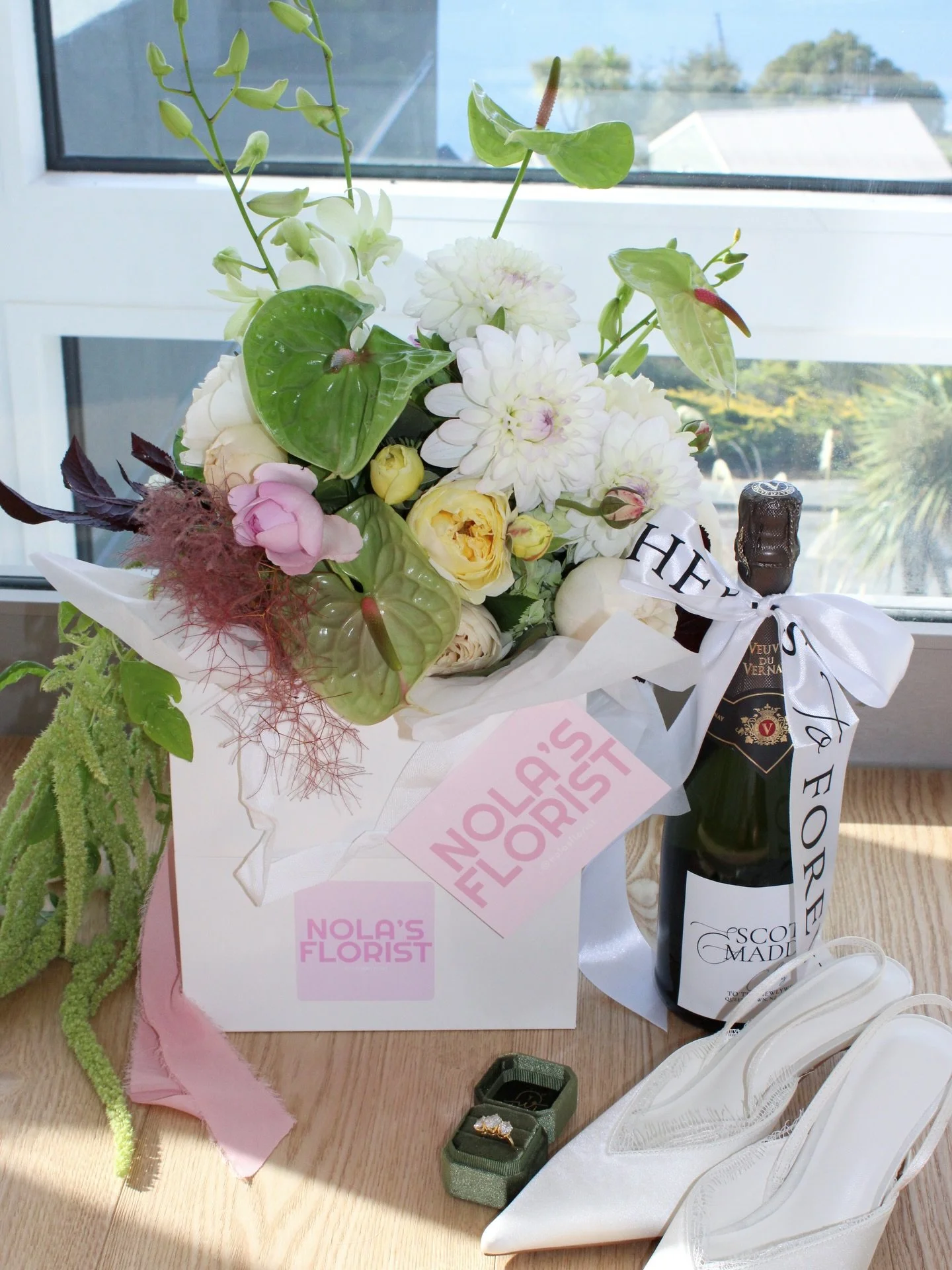 A bouquet of white, pink, and yellow flowers in a white box labeled "NOLA'S FLORIST", with a bottle of Veuve Clicquot champagne, a ring in a green jewelry box, and a pair of white shoes nearby, all set on a wooden surface by a window.
