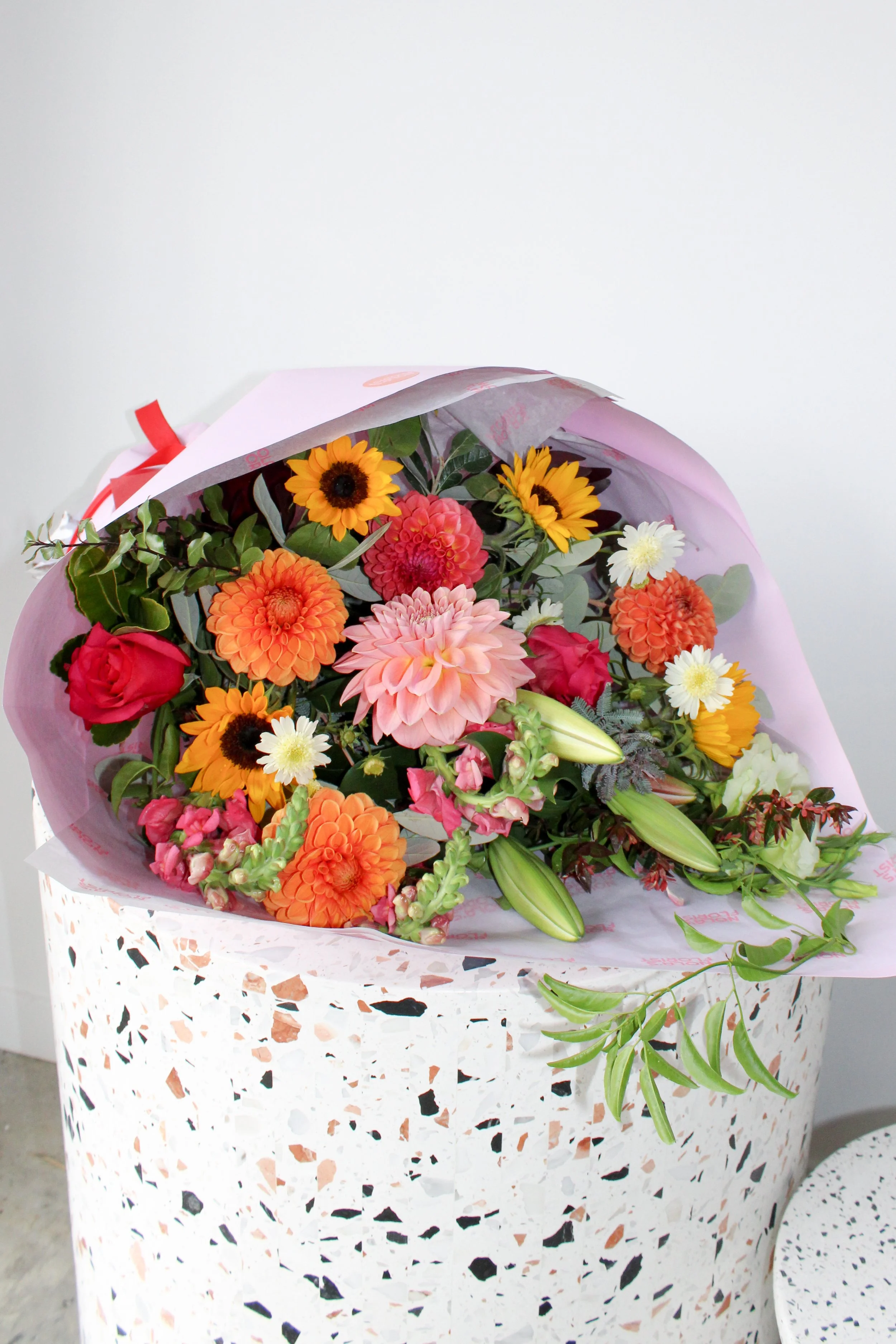 A bouquet of mixed flowers including pink dahlias, sunflowers, white daisies, orange dahlias, and red roses, wrapped in pink and white paper, placed on a terrazzo stand.