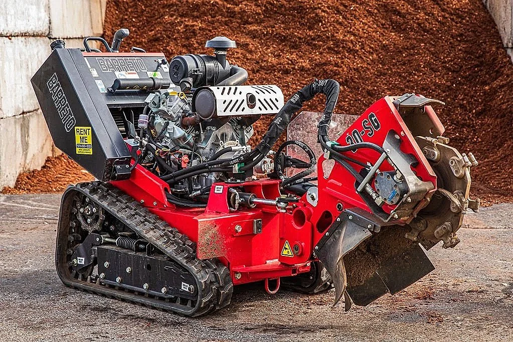 A red remote-controlled bulldozer with tracks, metal blade, and engine components, parked on pavement with dirt and concrete wall in background.
