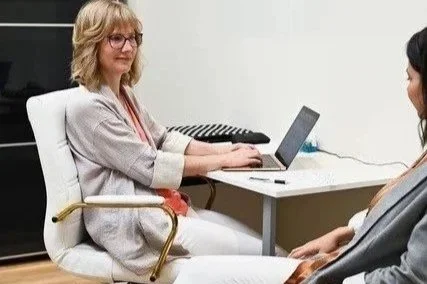 A woman in glasses sitting at a table using a laptop while talking to another person in an office setting.