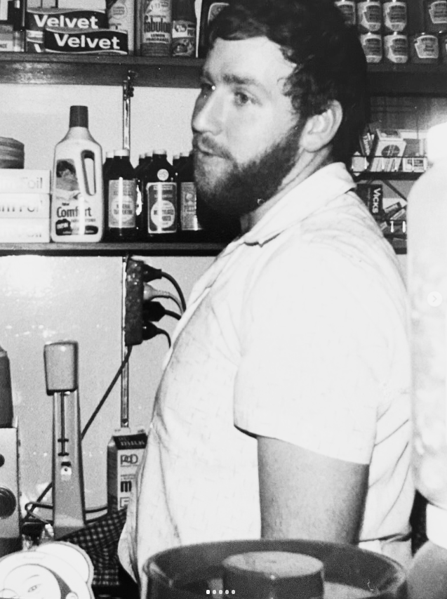 A man with dark hair and a beard wearing a light-colored shirt standing in front of shelves stocked with various bottles and cans in a store or workshop setting.