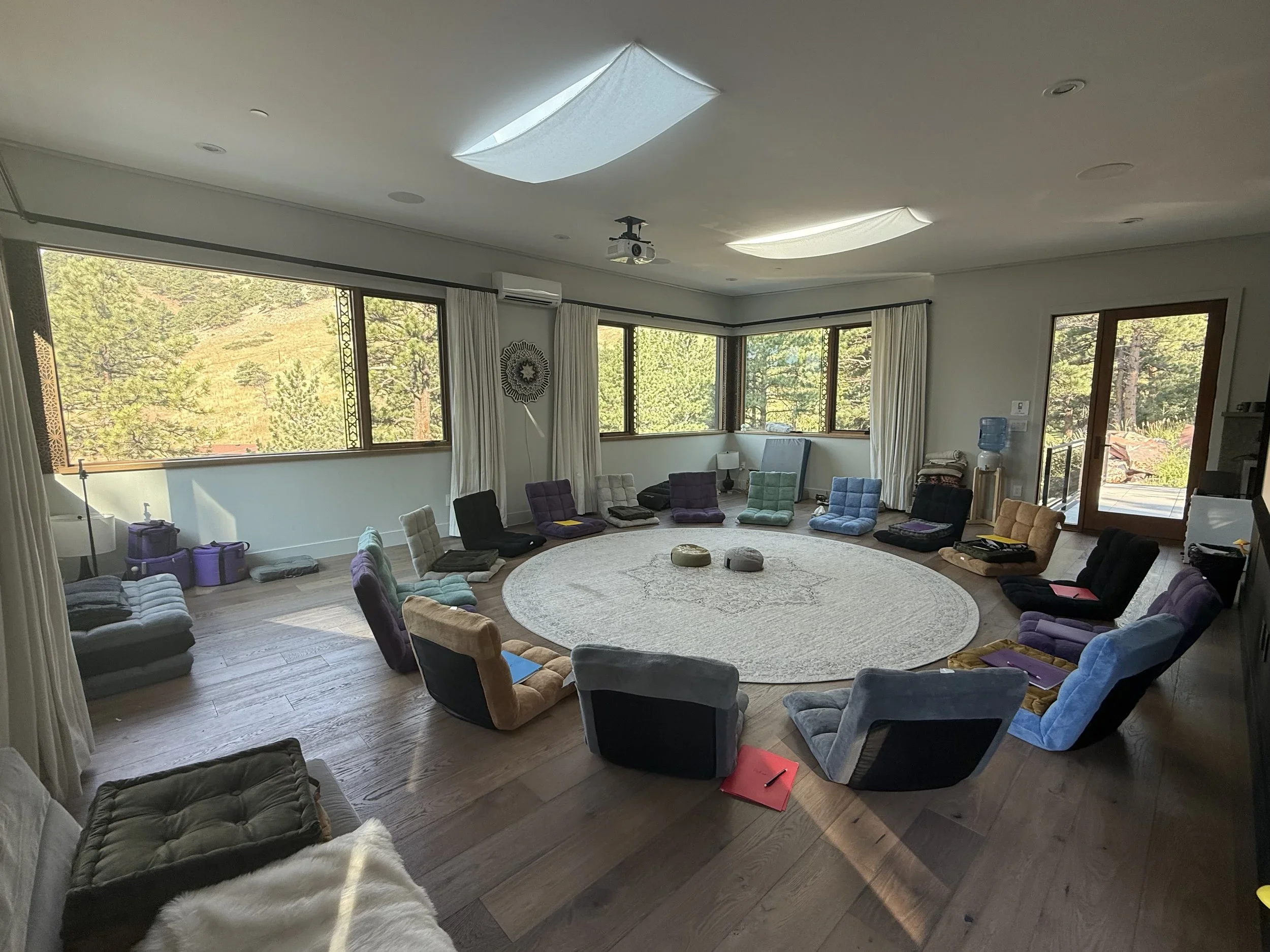 A round room with large windows surrounding it, filled with colorful floor chairs arranged in a circle, a light-colored rug in the center, and natural light coming from the windows.