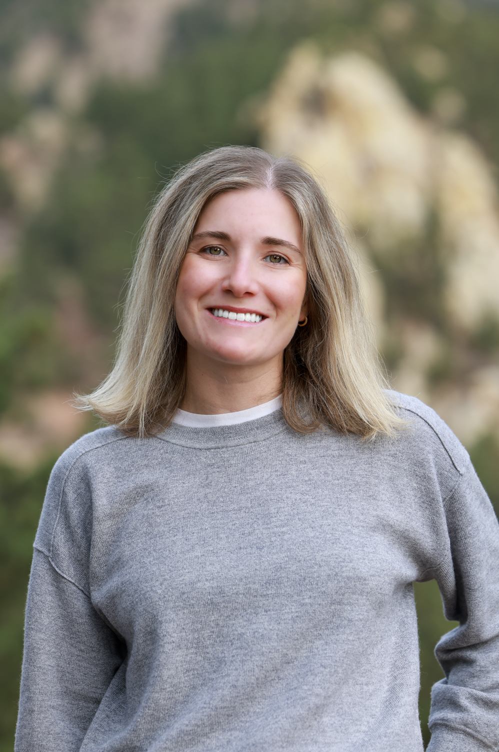 A woman with shoulder-length blonde hair, wearing a gray sweatshirt, smiling outdoors with a blurred natural background.
