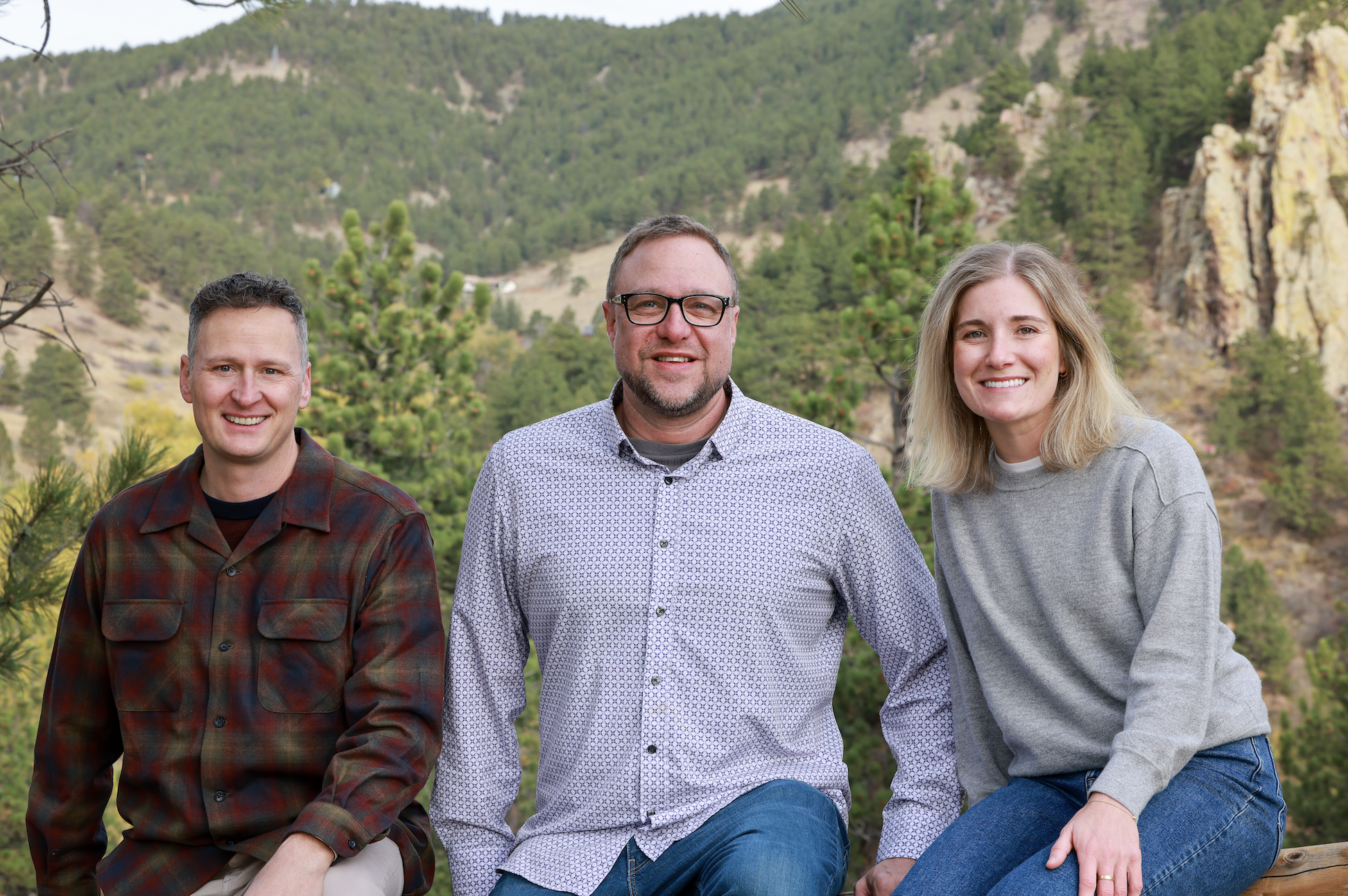 Three adults sitting outdoors with a mountainous, forested background, smiling at the camera.