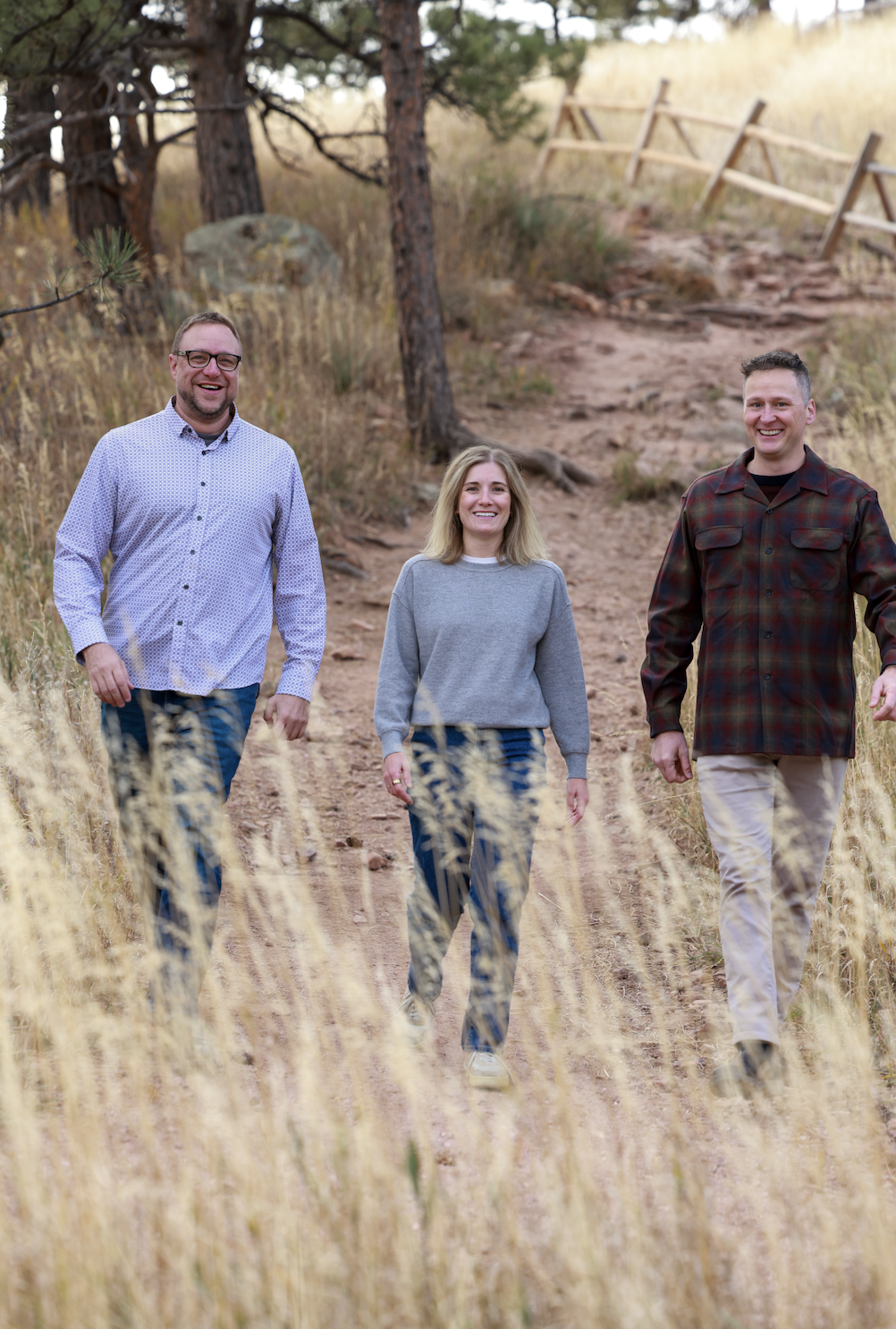 Three people walking on a dirt trail surrounded by dry grass, trees, and a wooden fence.