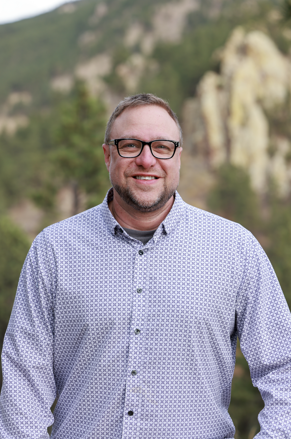 A man with glasses and a short beard smiling outdoors, wearing a patterned button-up shirt, with mountains and green trees in the background.