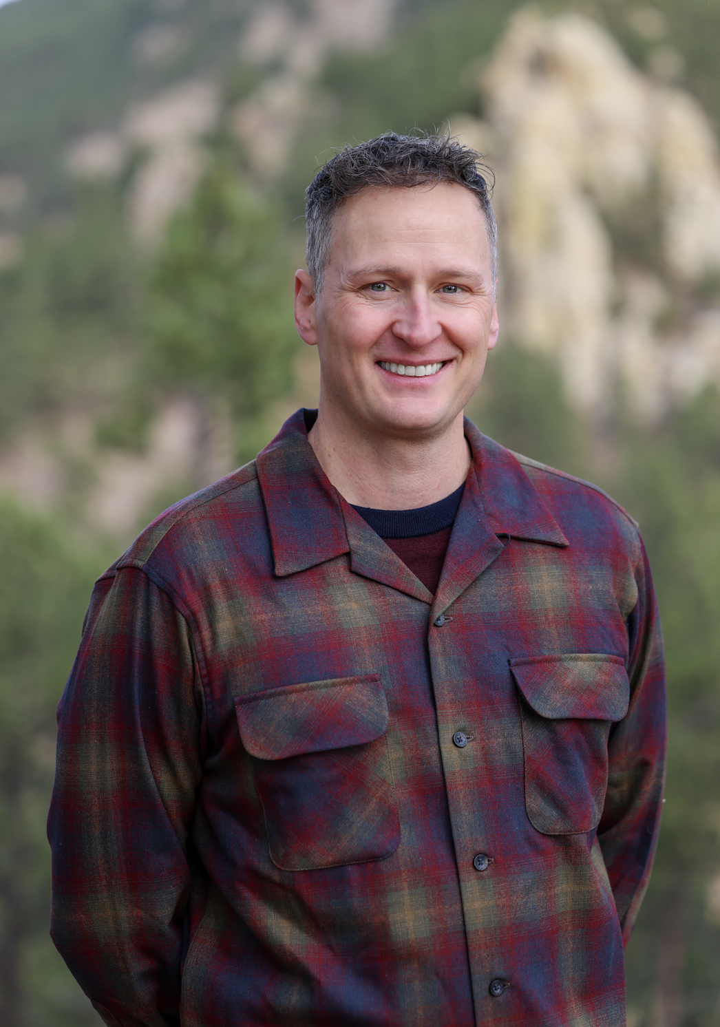 A man with short brown hair, wearing a red and green plaid jacket, smiling outdoors with trees and rocky formations in the background.