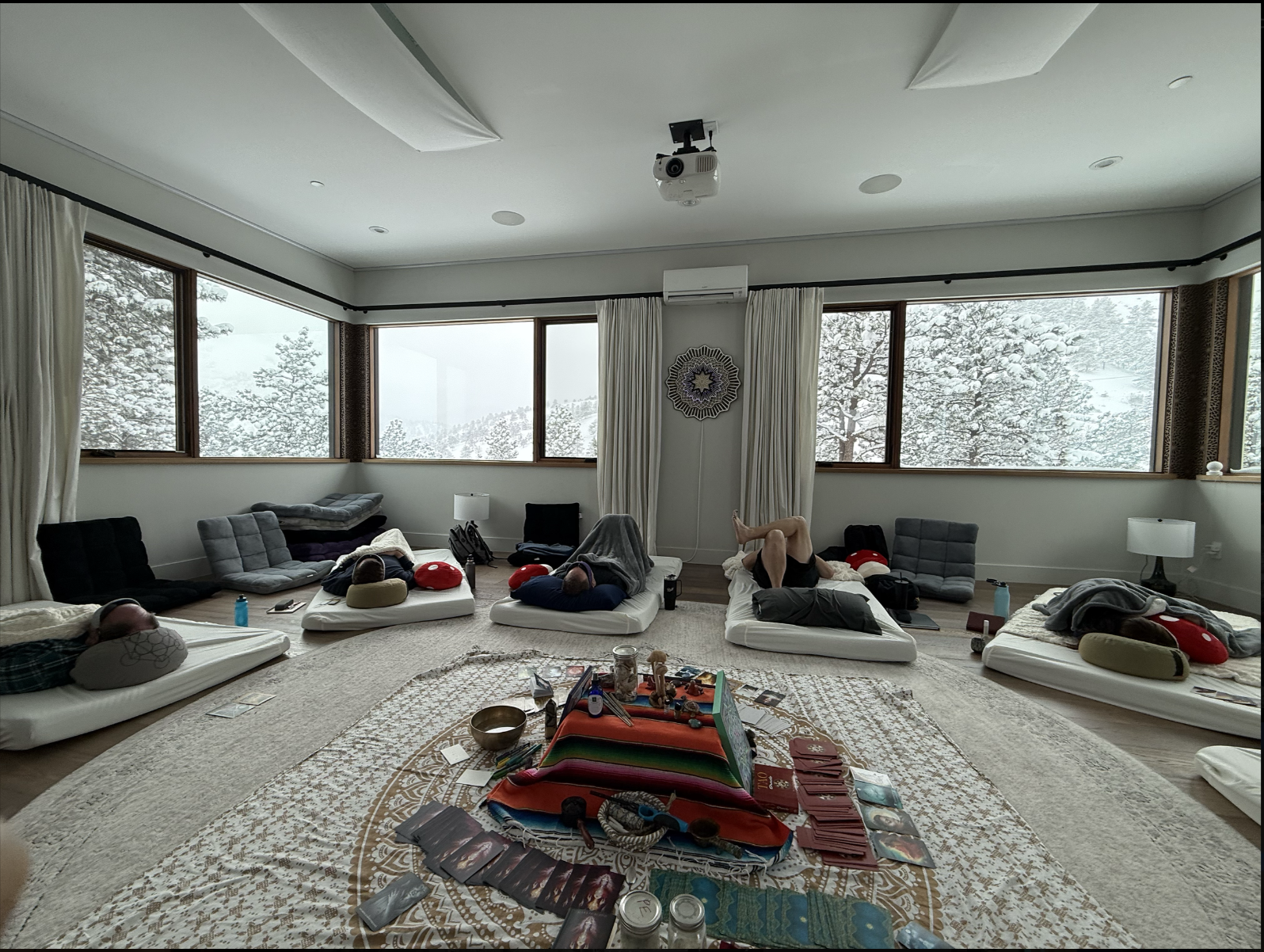 Group of five people lying on cushions and mattresses on a floor in a spacious living room with large windows showing snow-covered trees outside, surrounded by various objects including tarot cards, candles, and a blanket-covered table in the center.