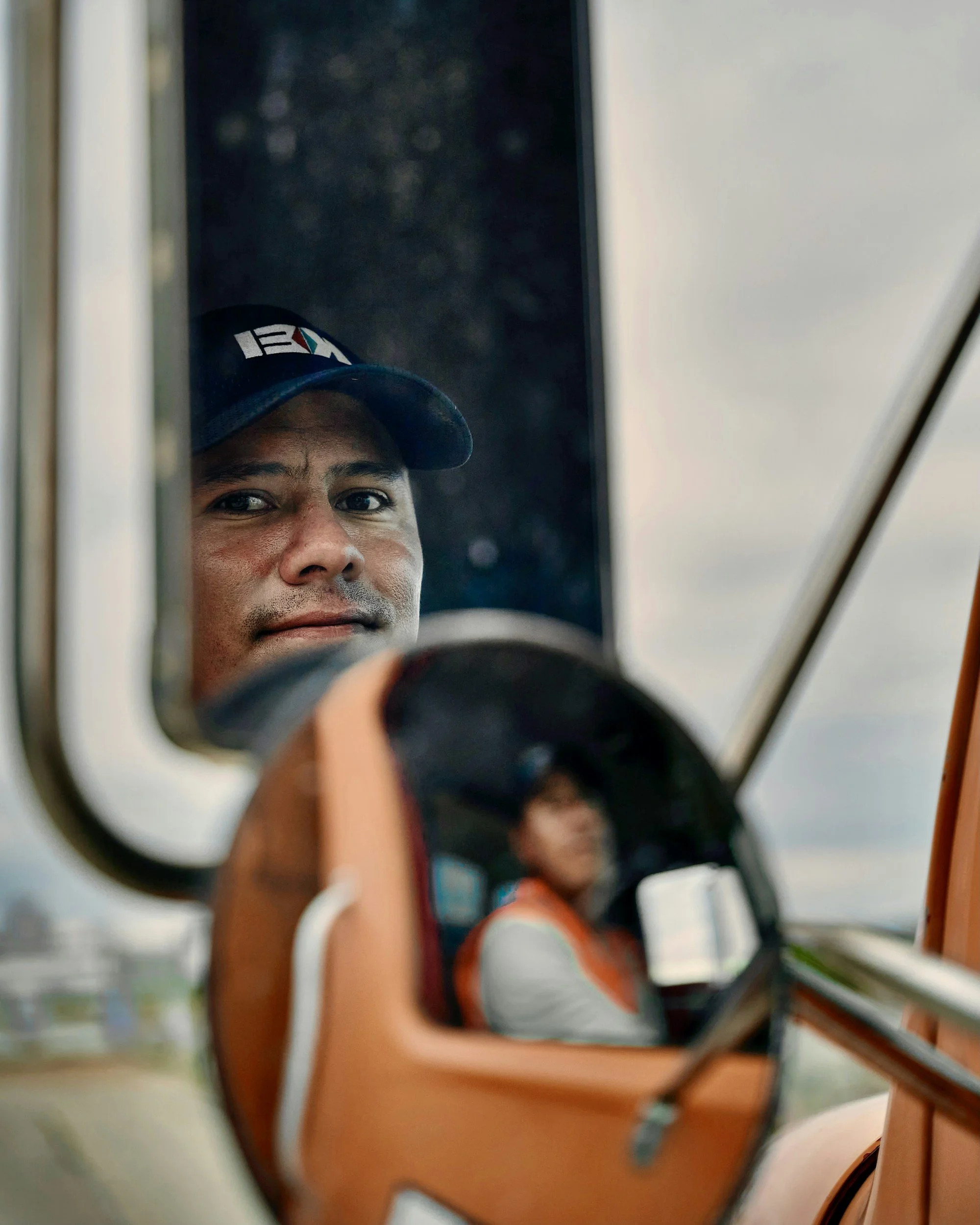 Close-up of a man's face reflected in a mirror, wearing a cap with a logo, holding a camera, there are blurred objects and a person in the background.