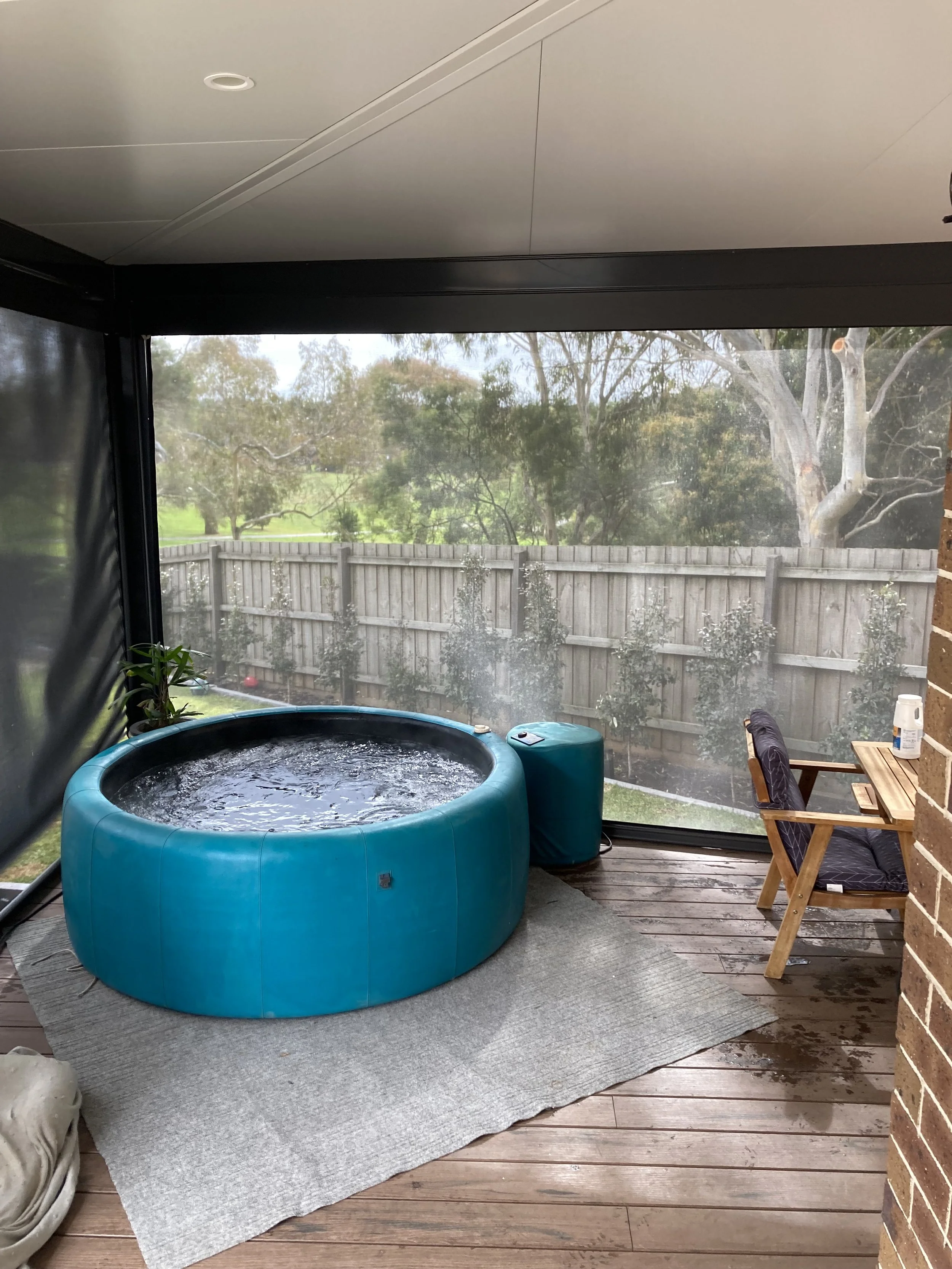 Outdoor screened porch with a blue hot tub, a small side table, two wooden chairs with cushions, and a bottle of sanitizer on a small table. Fenced backyard with trees and grass visible beyond the screen.