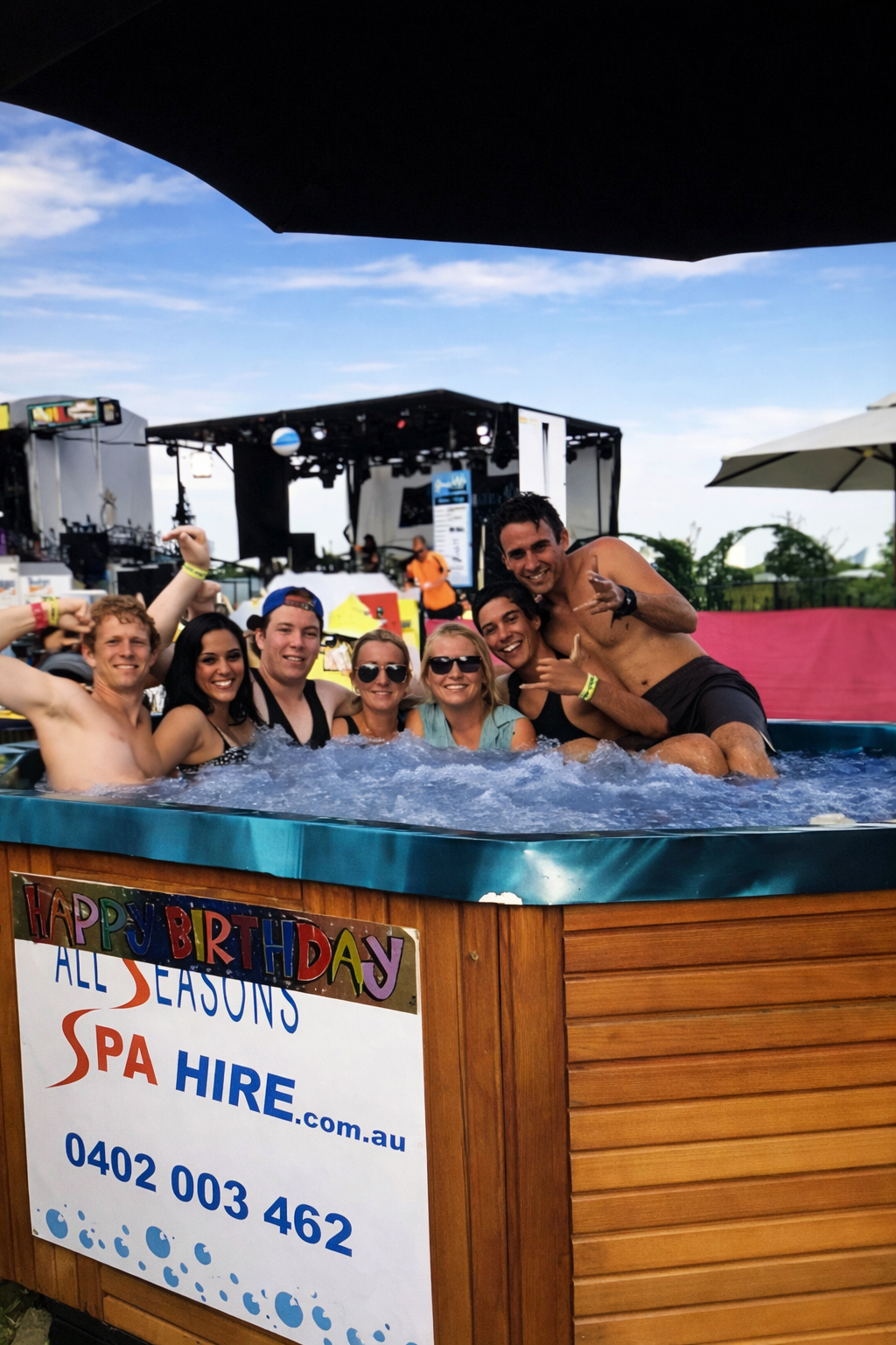 A group of seven young adults enjoying a birthday celebration in an outdoor hot tub at a festival, with a stage and festival tents in the background, under a partly cloudy sky.