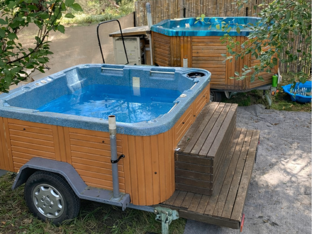 Two wooden hot tubs with blue interiors in an outdoor backyard setting, one on a wooden deck with steps, surrounded by trees and greenery.