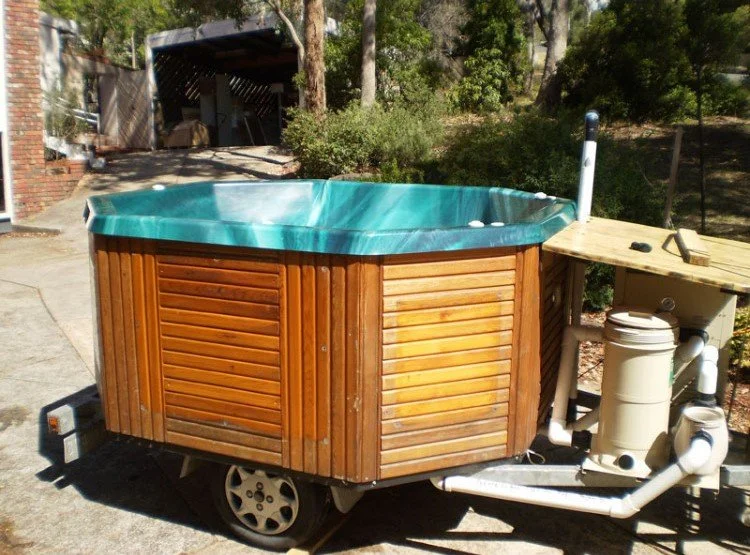A wooden hot tub on a trailer with a green interior, situated outdoors on concrete with trees and a brick building in the background.