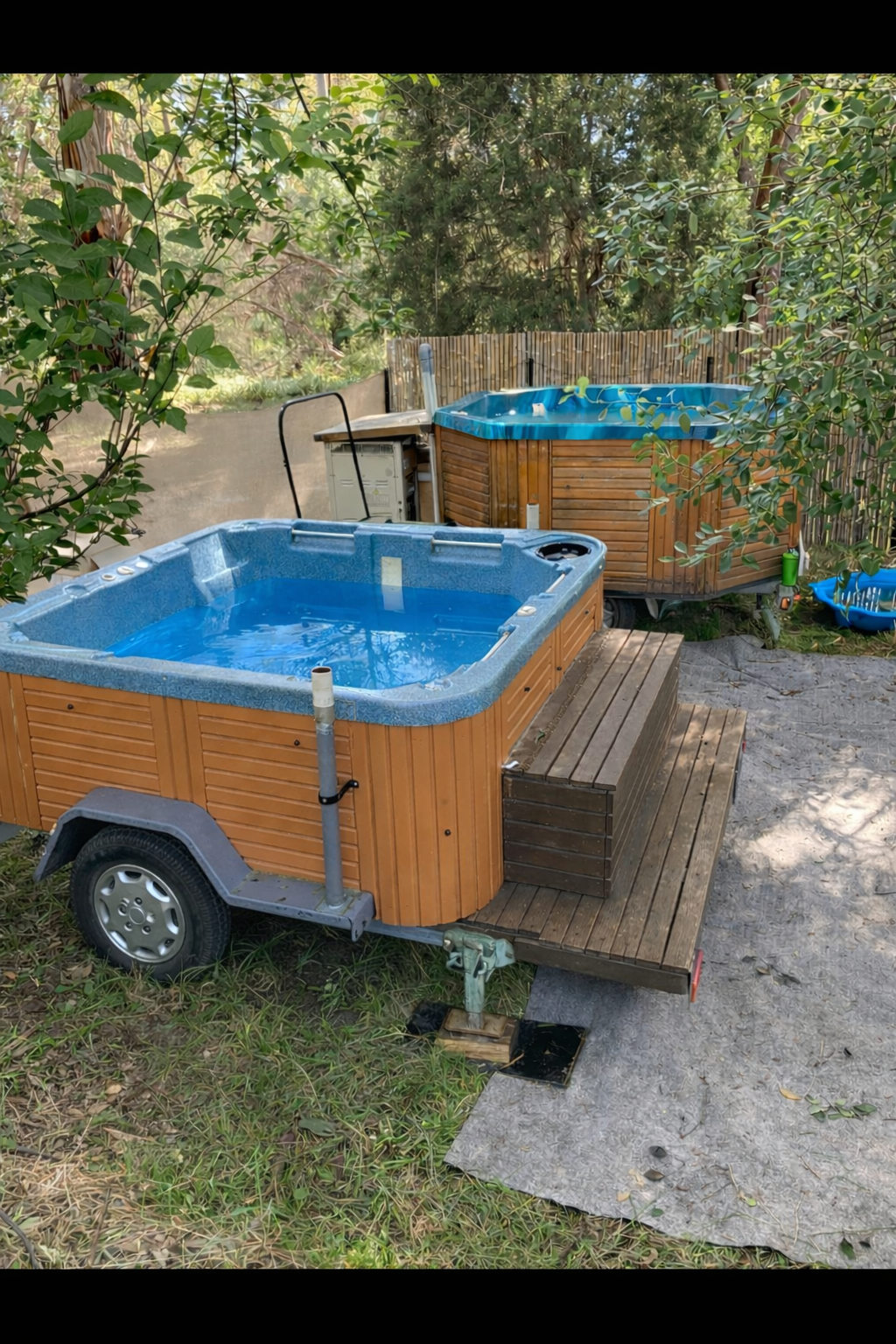 Two hot tubs, one on a trailer and one in the background, in a backyard with trees and plants, surrounded by a bamboo fence.