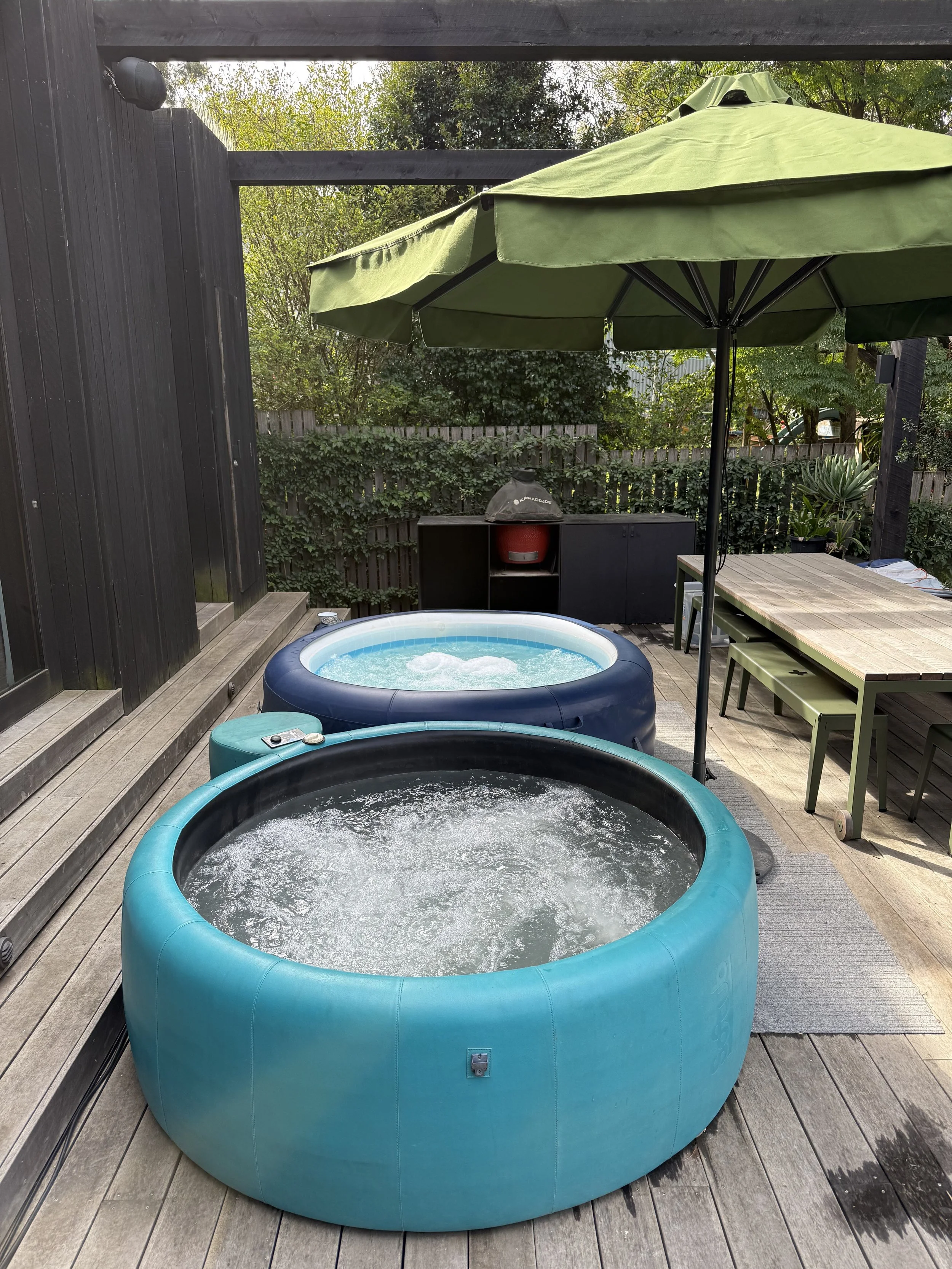 A backyard patio with two hot tubs, one blue and one black, placed on a wooden deck. There is a large green patio umbrella shading part of the area, a barbecue grill, and outdoor furniture including a table with a bench and chairs. A wooden fence and greenery are in the background.