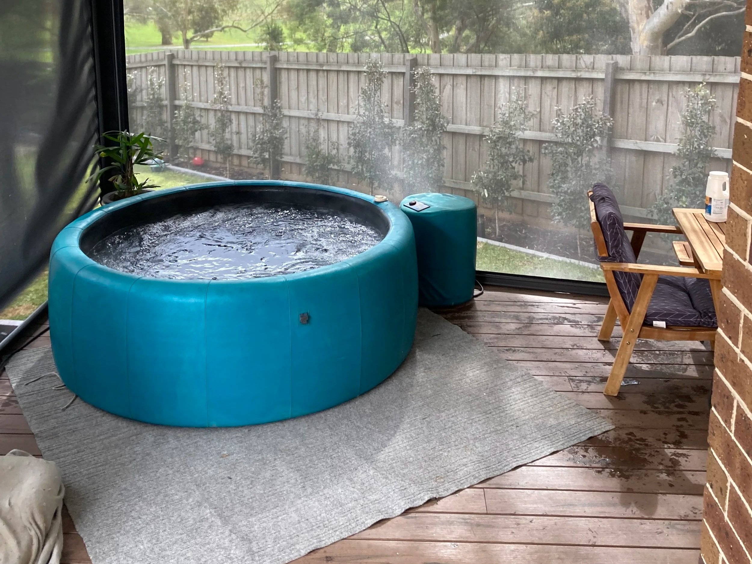 An outdoor patio area with a blue hot tub filled with water, a plant in a corner, a small wooden table with a chair, and a large window revealing a fenced backyard with trees and grass.