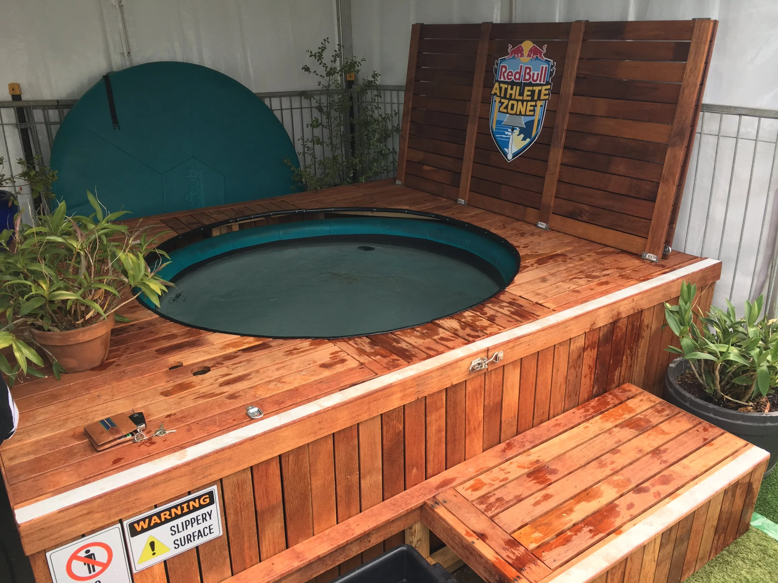 A hot tub with a wooden surrounding structure, covered by a wooden shield with a Red Bull Athletes Zone sign, filled with water and placed in an indoor or sheltered environment. There are potted plants nearby, and keys and a wallet on the wooden surface.