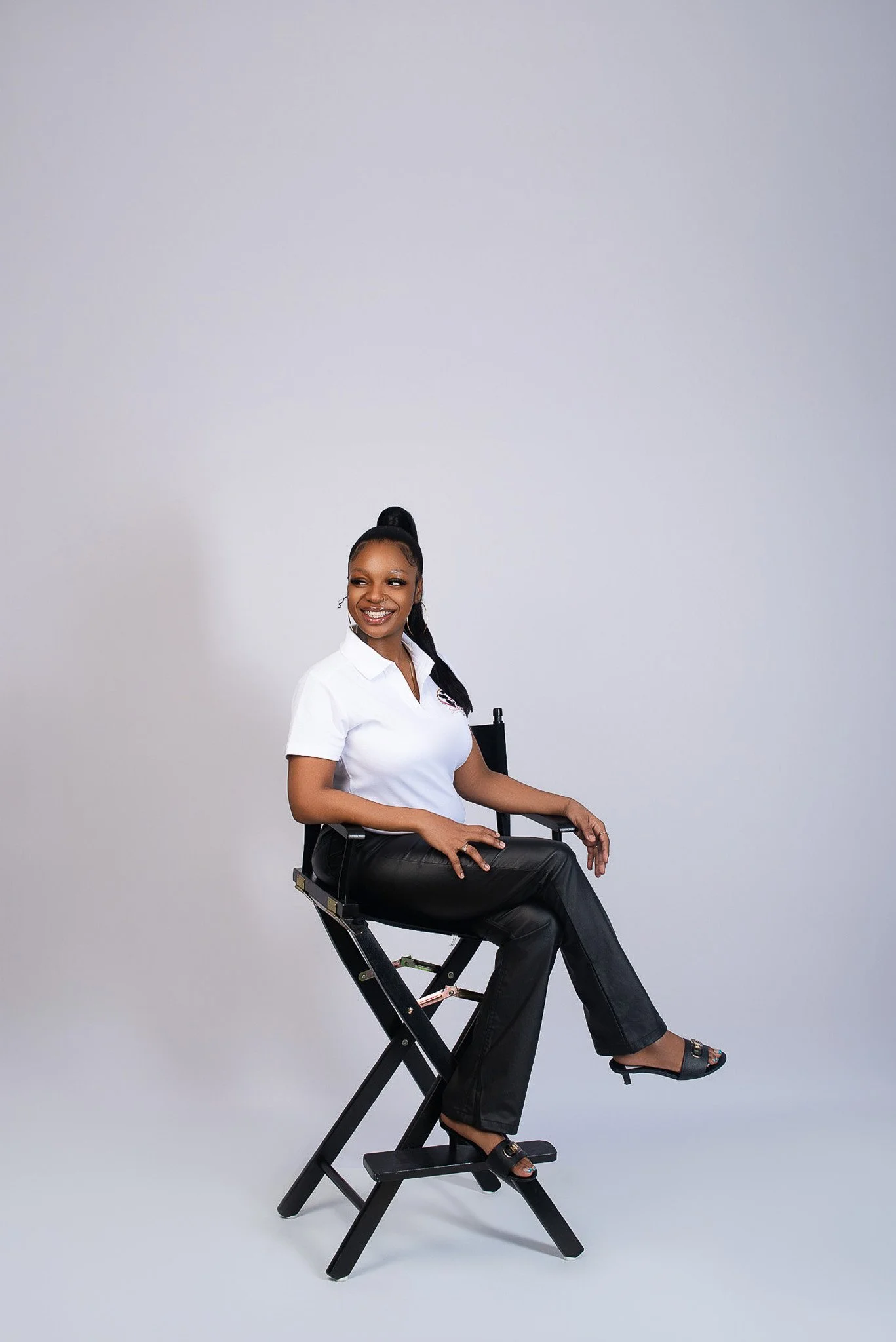 A woman with dark hair styled in a high ponytail sitting on a director's chair against a plain, light-colored background. She is smiling and wearing a white polo shirt, black leather pants, and open-toed shoes.