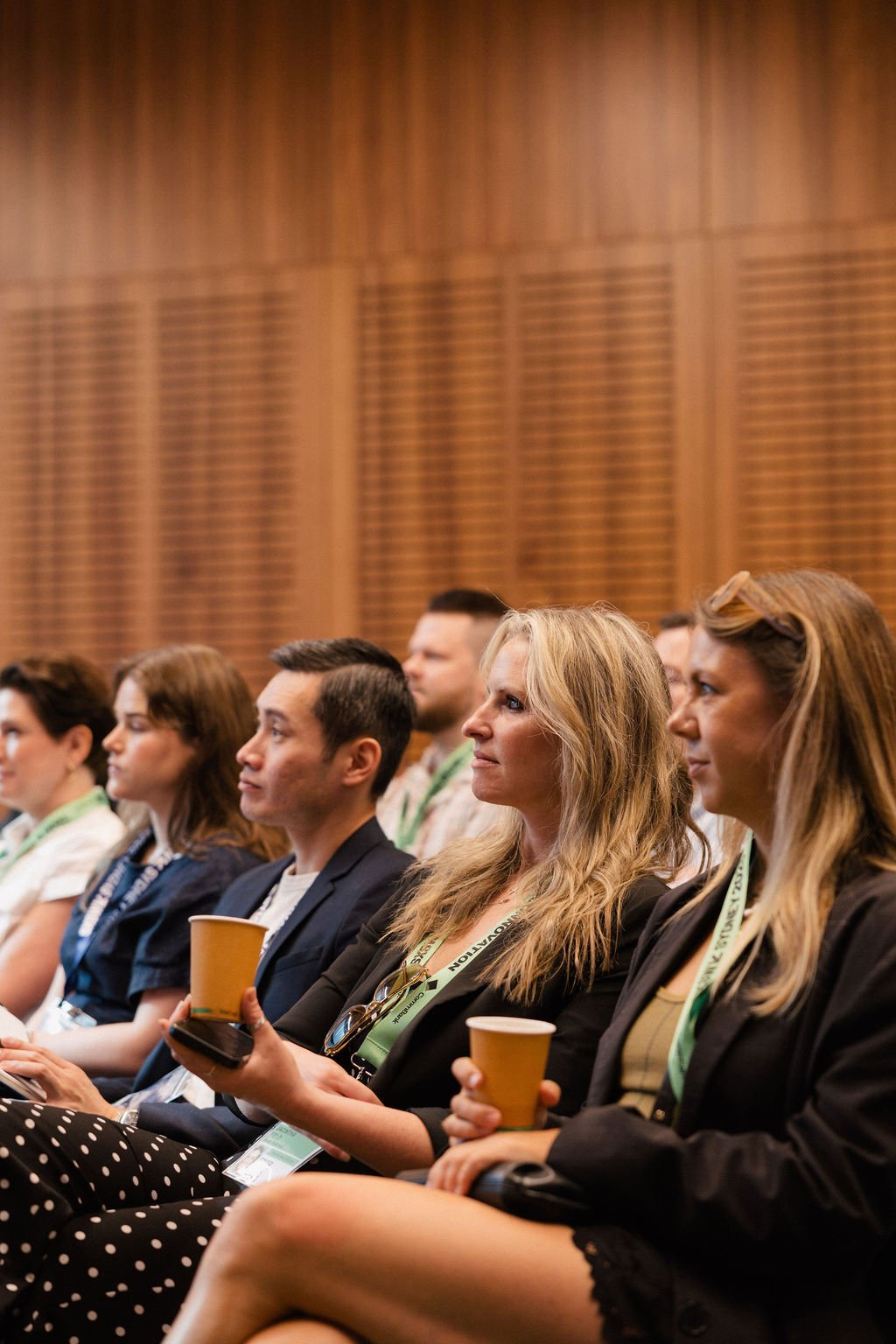 People attending the Impact Stage in 2025, sitting in a row, holding cups, listening attentively, with wooden panel background.