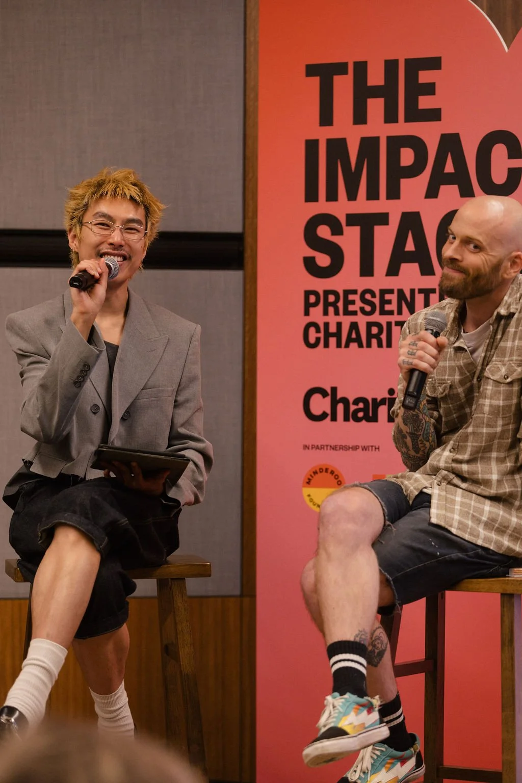 Two men sitting on stools speaking into microphones at a charity event, with a pink banner behind them reading 'The Impact Stage Presents Charity'.