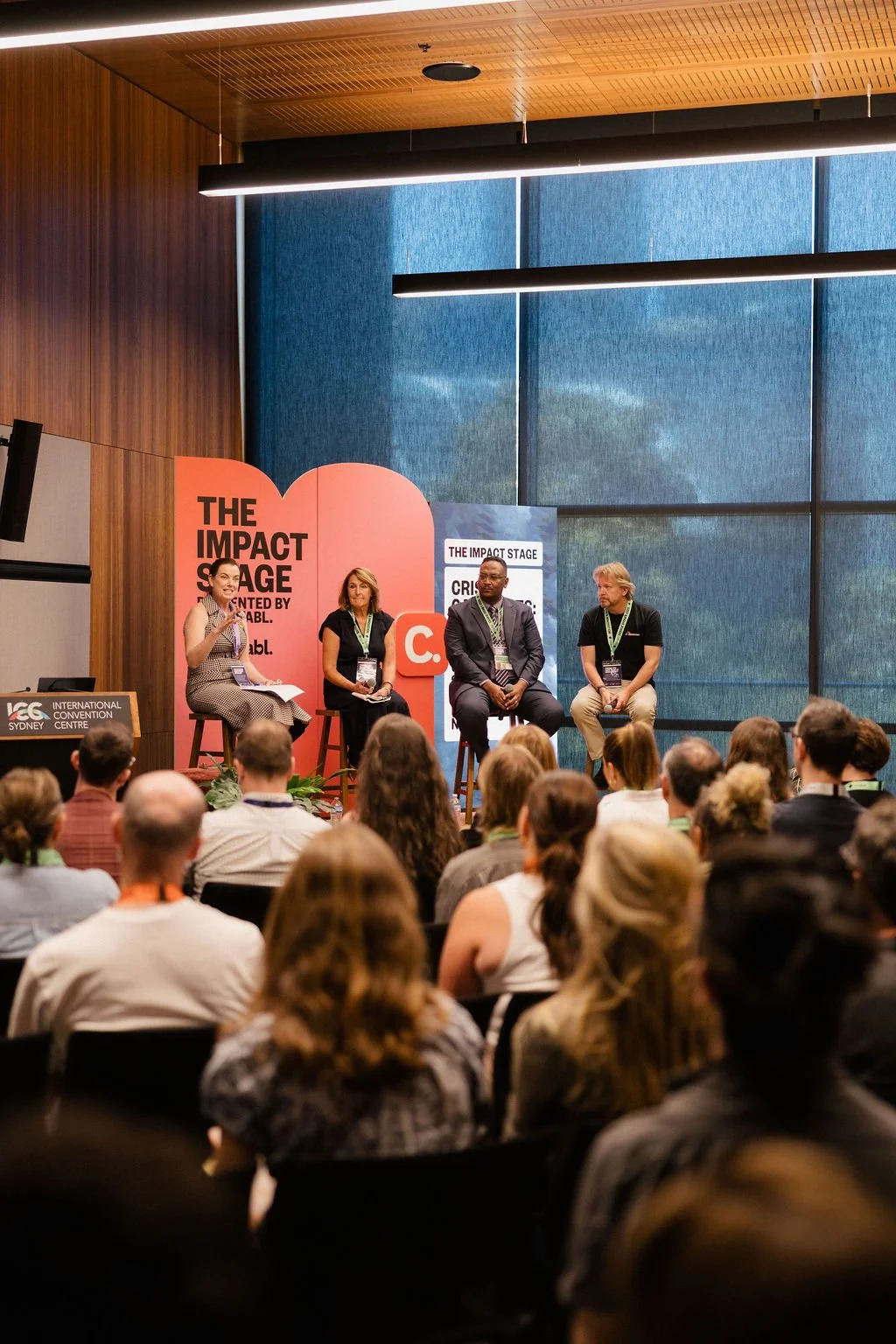 Four panelists speaking on stage at a conference, seated on stools with one woman gesturing at the Impact Stage 2025 presented by Charitabl.