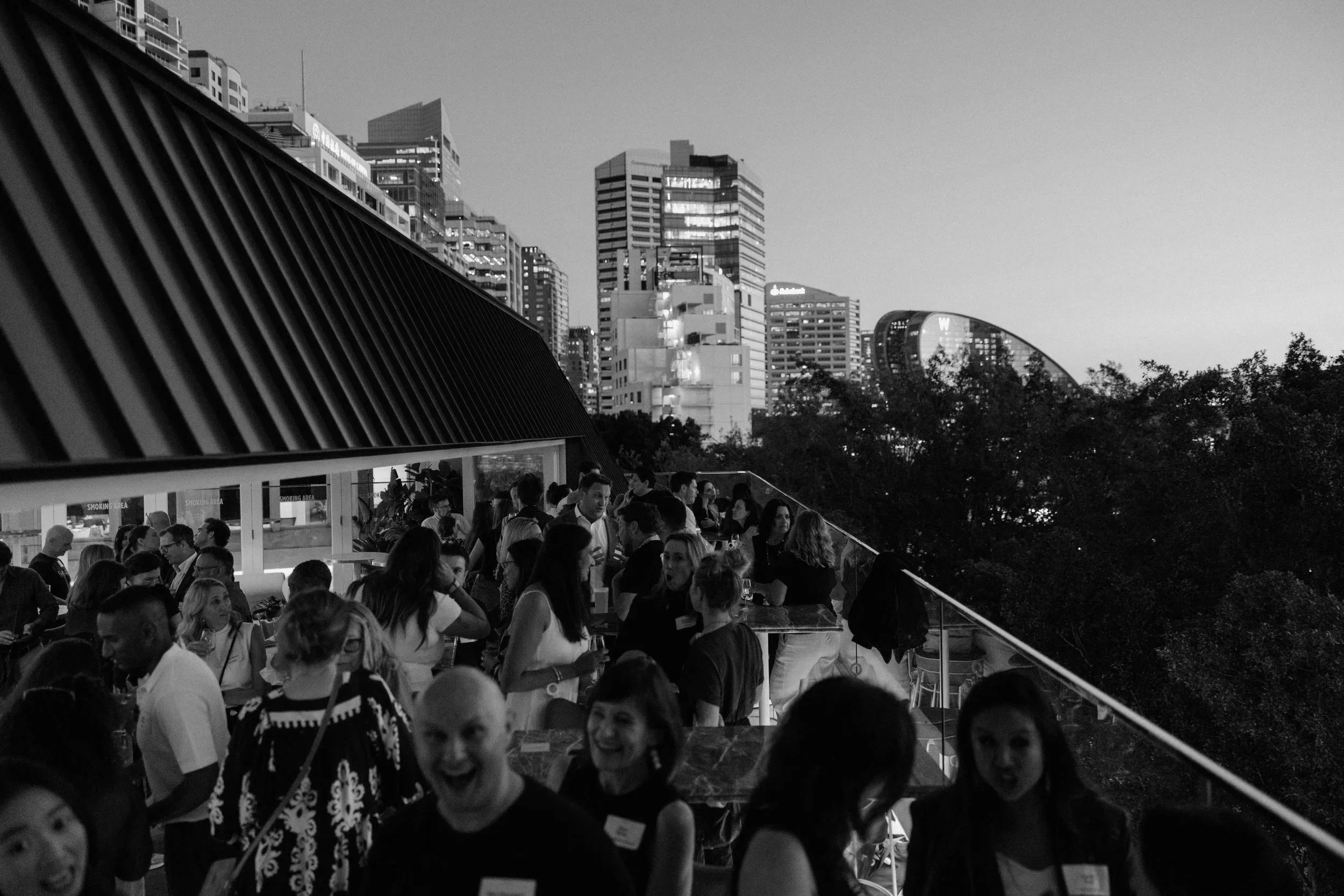 People socializing on a rooftop terrace with city skyline in the background, viewed in black and white.