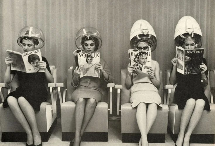 Four women sitting under hair dryers at a beauty salon, each reading a different magazine.
