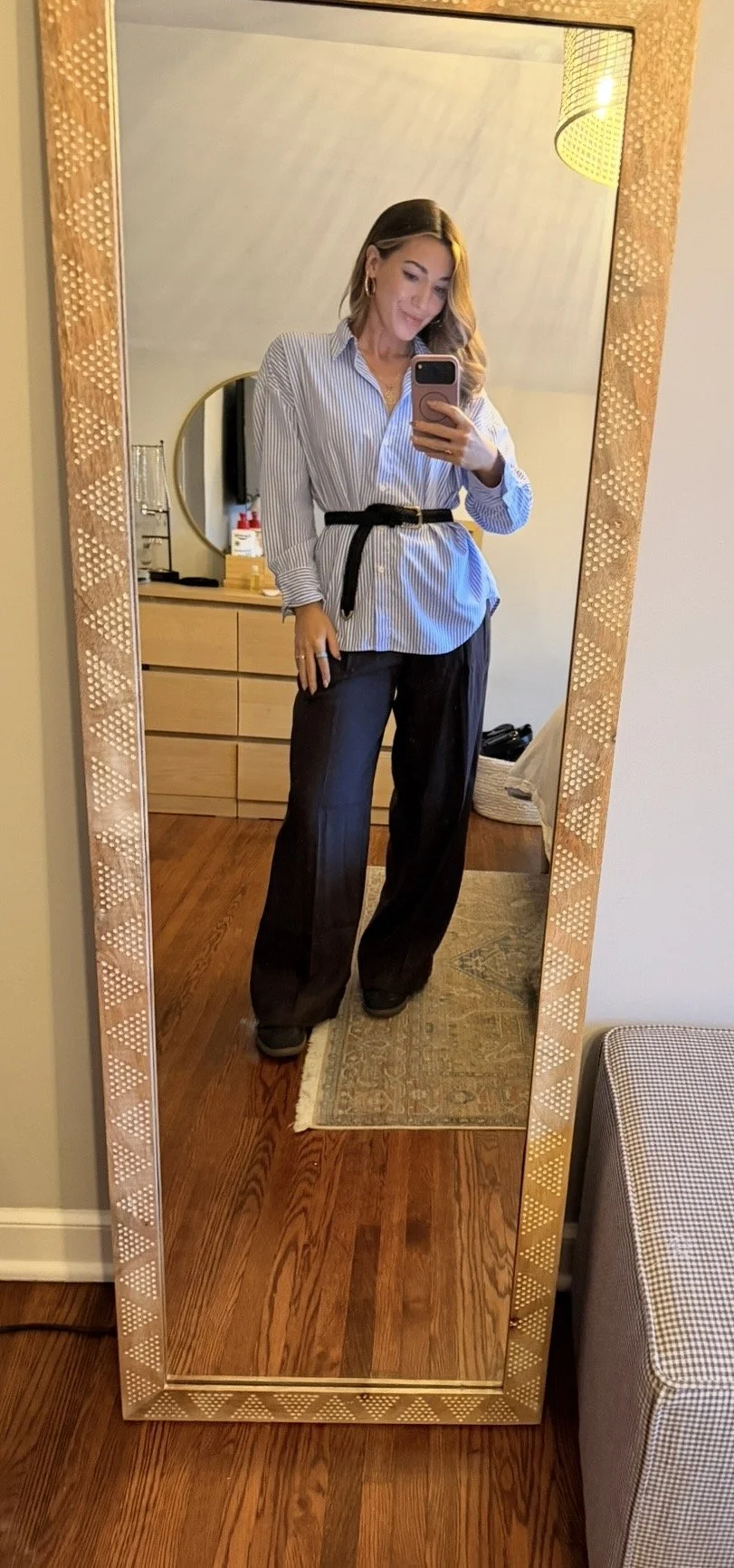 A young woman taking a mirror selfie in a room with wooden flooring, wearing a striped button-up shirt tied at the waist, black pants, and high heels.