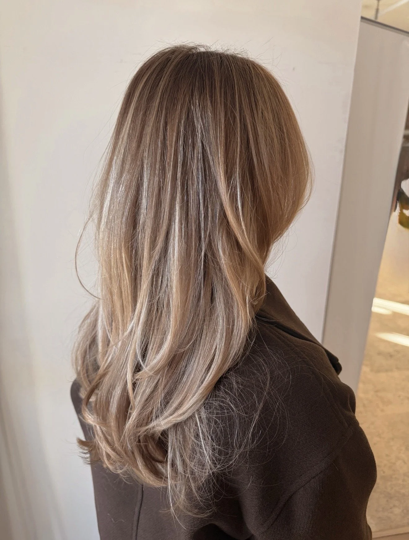 Side view of a woman with long, wavy, blonde hair wearing a dark brown top, standing against a light-colored wall.