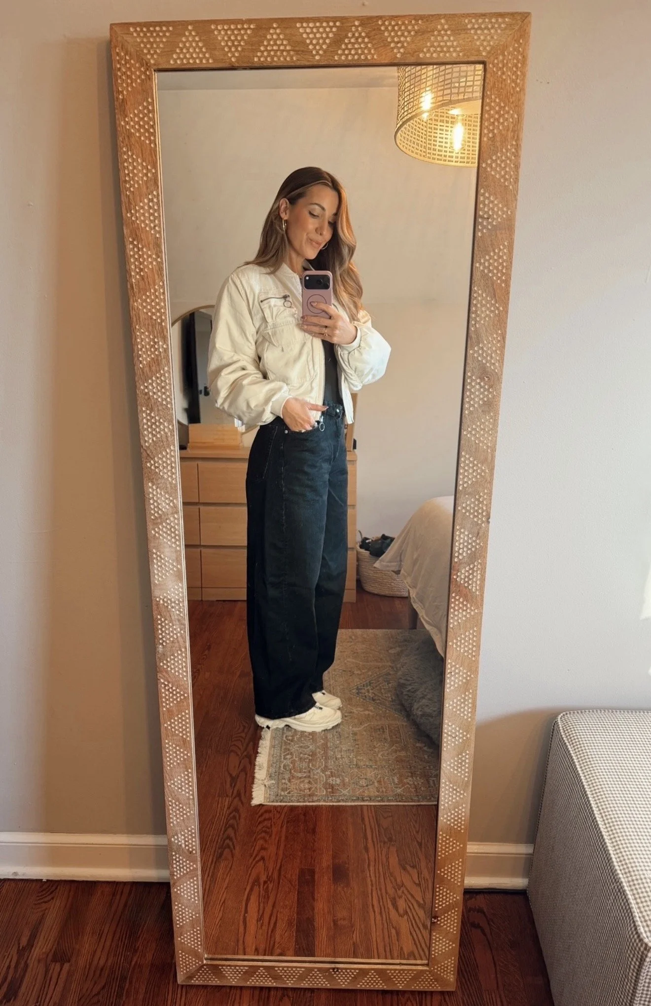 A woman taking a mirror selfie shows her wearing a cream jacket, black high-waisted pants, and sneakers in a room with wooden flooring and beige walls.