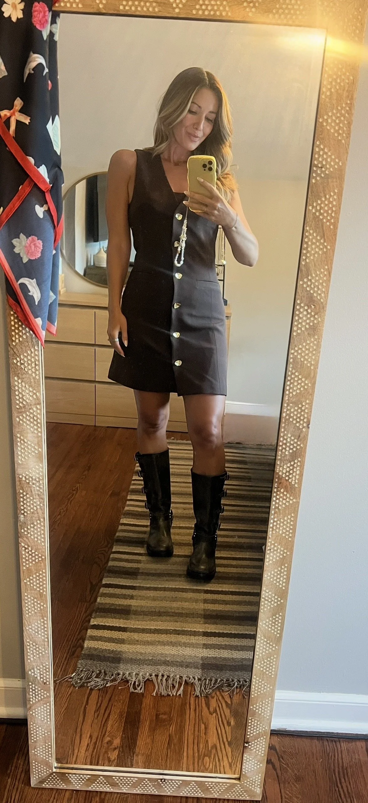 A woman taking a mirror selfie, wearing a black sleeveless dress with buttons down the front and black knee-high boots.