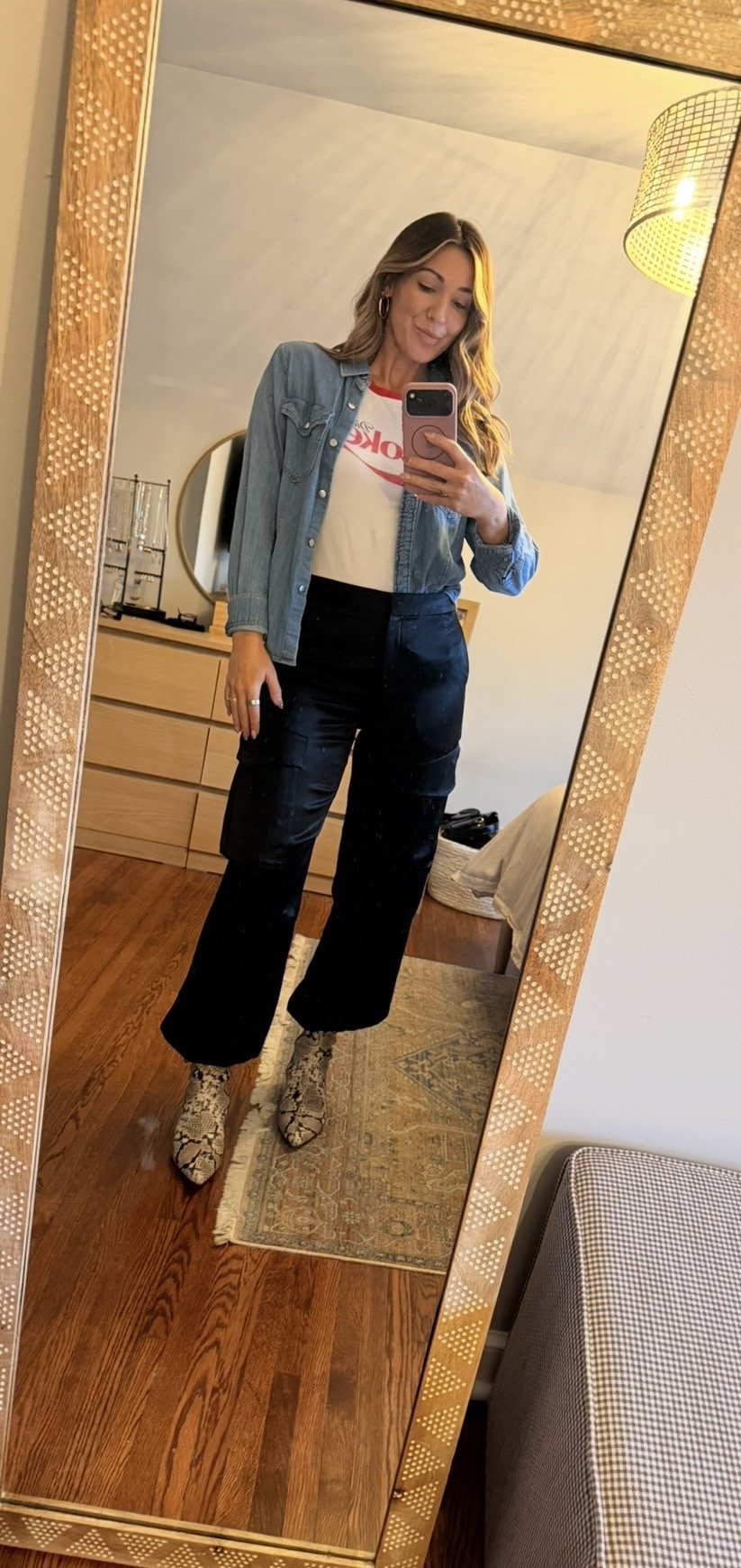 A woman taking a mirror selfie in a bedroom, wearing black wide-leg pants, a white T-shirt with red lettering, a denim jacket, and snake print boots.