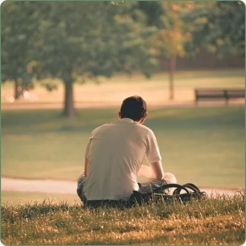 A person sitting on grass in a park with a backpack, looking at the scenery with trees and a park bench in the background.