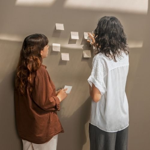Two women with long hair standing in front of a gray wall with sticky notes attached, discussing and pointing at the notes.
