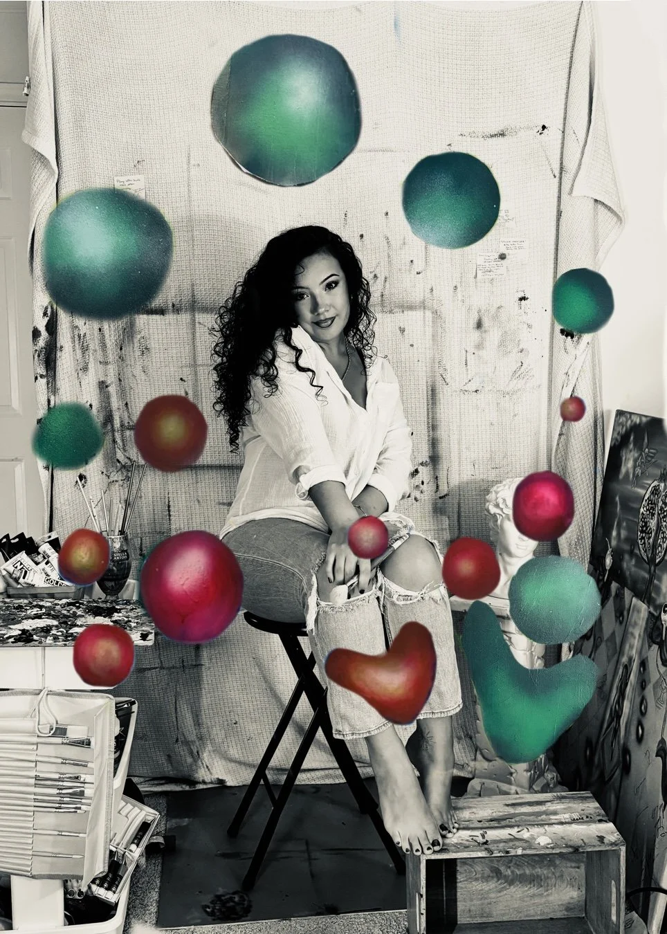 Black and white photo of a woman sitting on a stool in an artist's studio, surrounded by floating colorful paint splatters and spheres, with art supplies and a sculpture in the background.