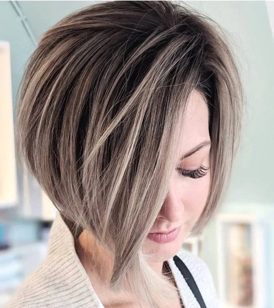 A woman with a stylish, layered bob haircut with blonde and brunette streaks, leaning her head downward with her eyes closed.