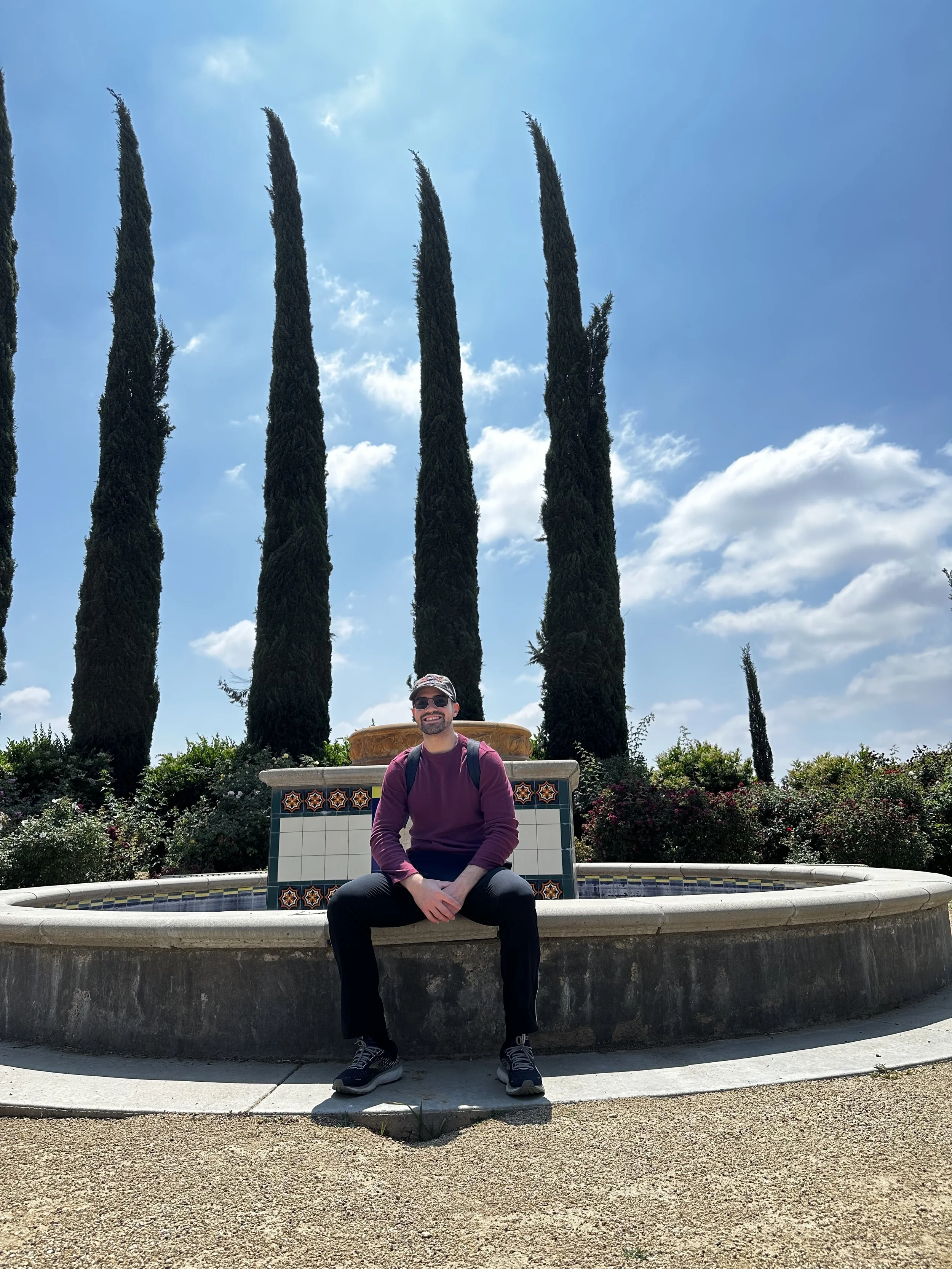 A man sitting on the edge of a fountain with tall cypress trees and a partly cloudy sky in the background.