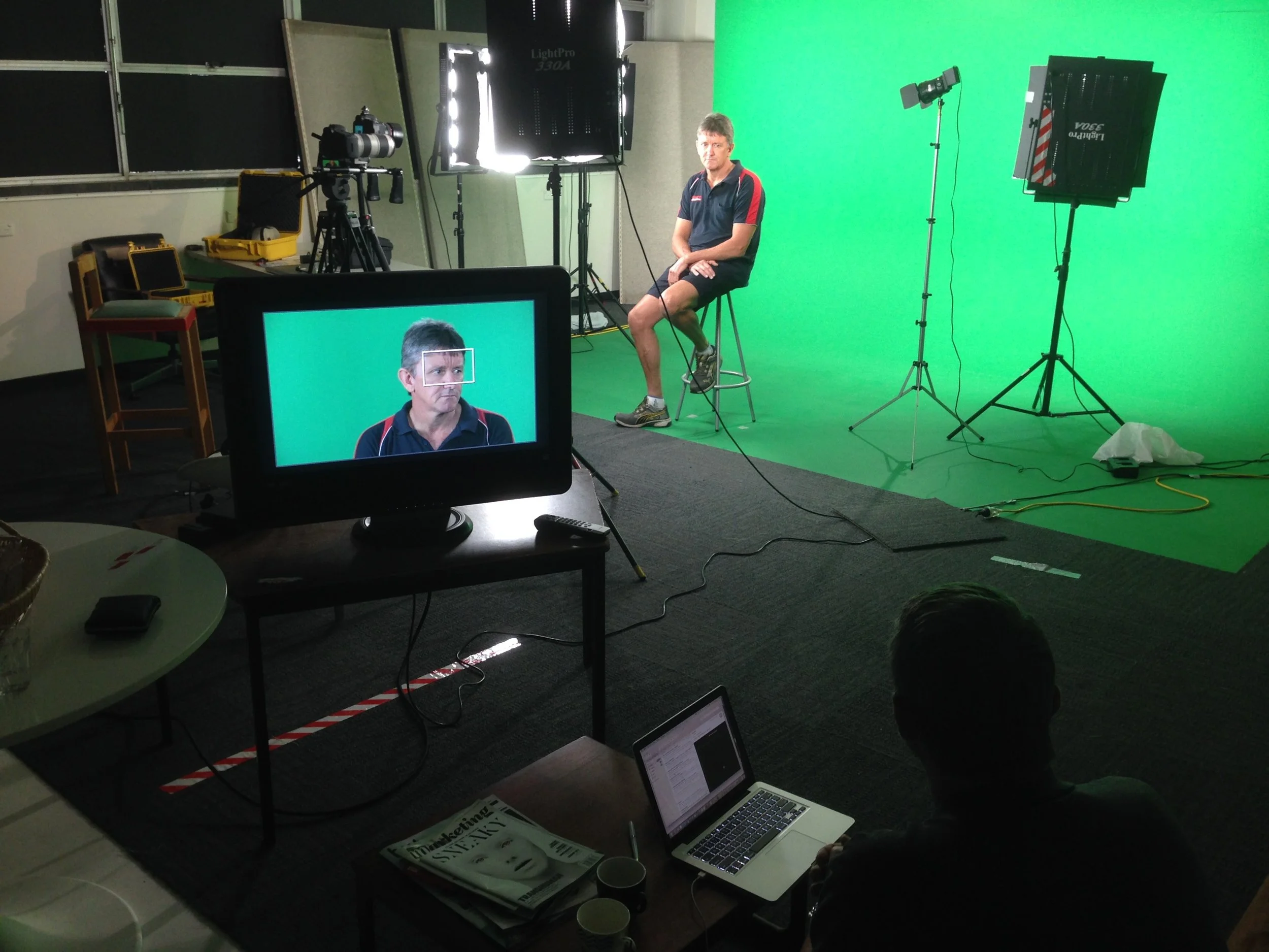 A video production studio with green screen and professional lighting. A man is seated on a stool in front of the green screen, preparing to be filmed. Equipment including cameras, lights, and monitors are set up around him.