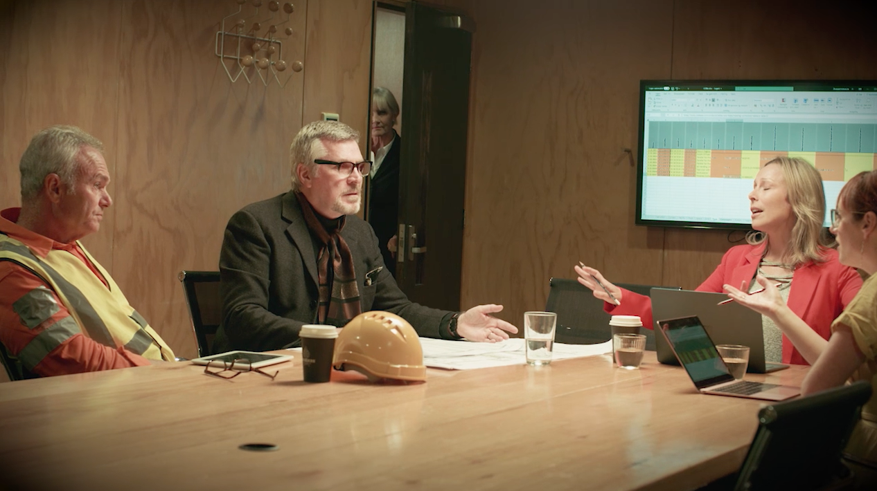 A group of five people having a meeting around a large wooden conference table in a room with wood-paneled walls. One woman in a red blazer appears to be speaking, while another woman in yellow listens. A man with glasses and a dark jacket is gesturing, and two men, one in a safety vest, are listening. There are glasses of water, a coffee cup, and a helmet on the table. A large monitor on the wall displays a colorful spreadsheet.