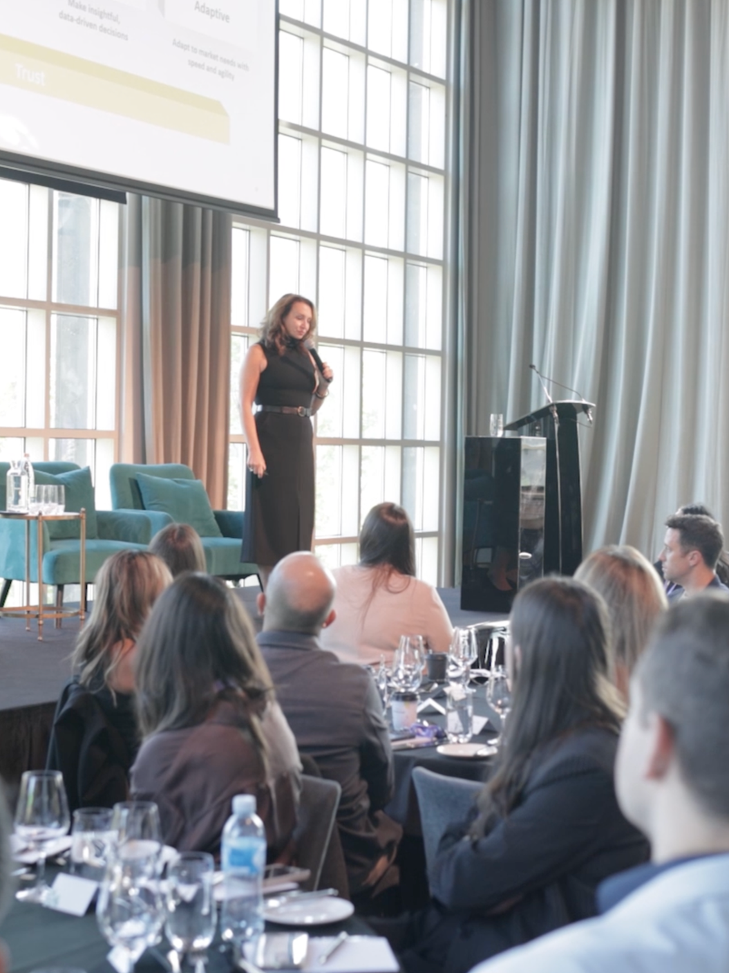 A woman in a black dress is giving a presentation on a stage in front of a seated audience in a conference room with large windows and curtains.