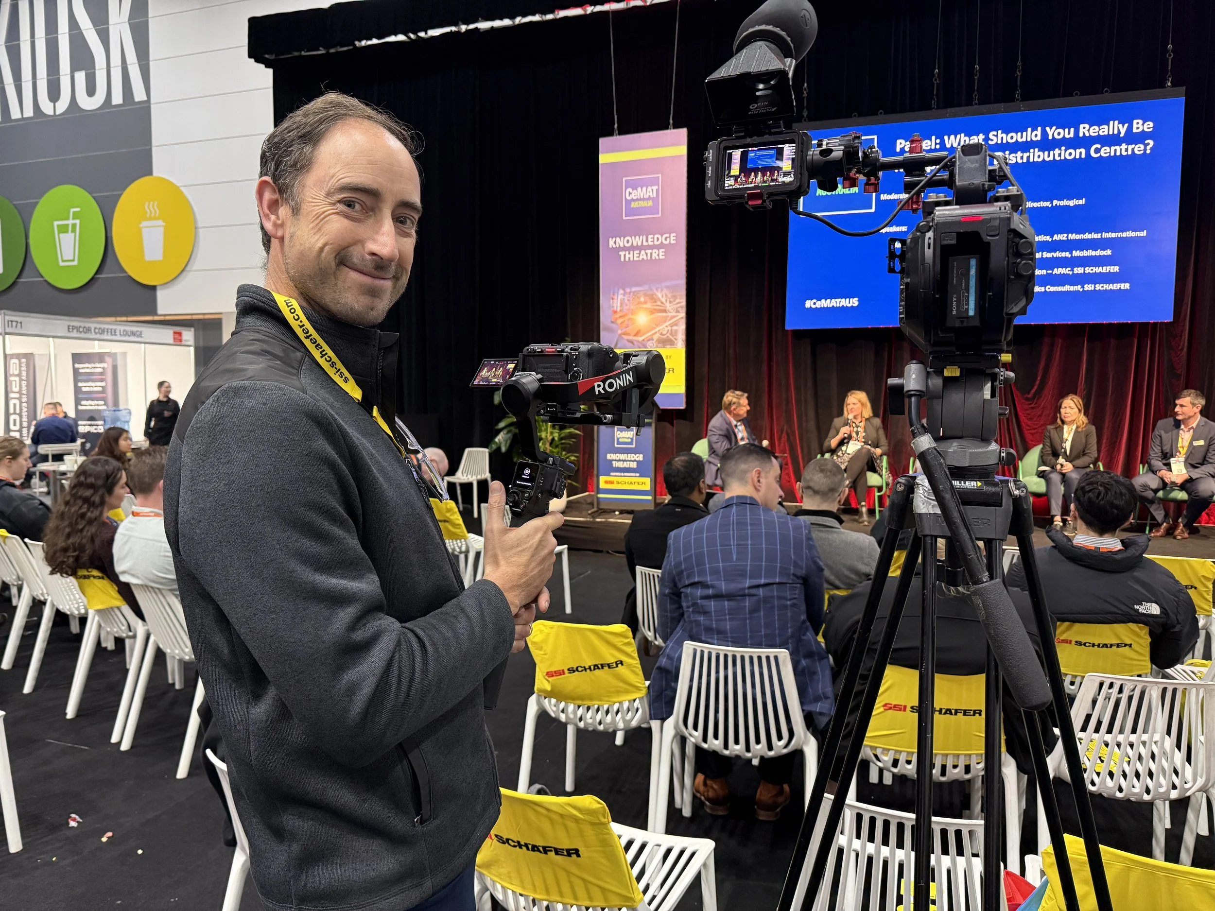 A man with dark hair and a beard in a gray jacket holding a professional camera captures a panel discussion at an event. The panel is seated on stage with a large blue screen behind them displaying the title of the discussion. Attendees are seated in white chairs, some with yellow bags, and promotional banners and signs are visible in the background.