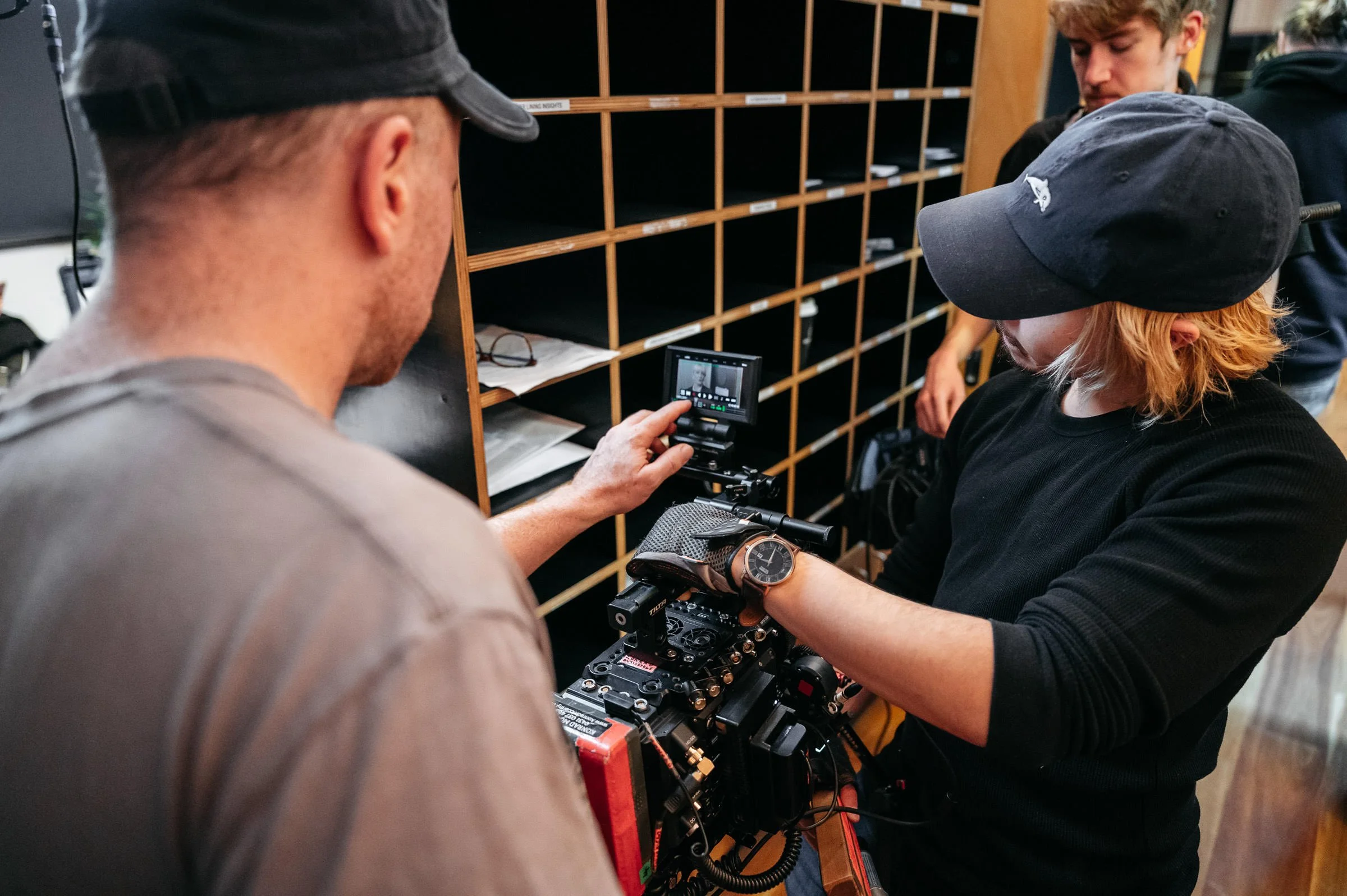 A person with a watch on their wrist is operating a camera rig, with two other people inspecting or adjusting equipment nearby in an indoor setting.