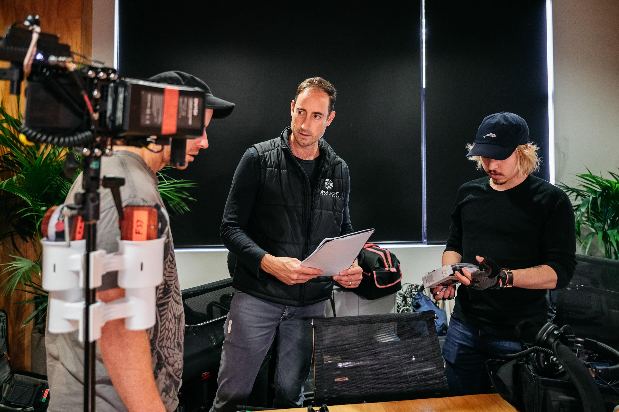 A film crew is gathered in a room with black walls and large windows covered with blinds. There are three men, one holding papers, another examining a device, and the third talking to the man with papers. Audio and video equipment is visible, including a camera on a tripod, and there are green plants in the background.