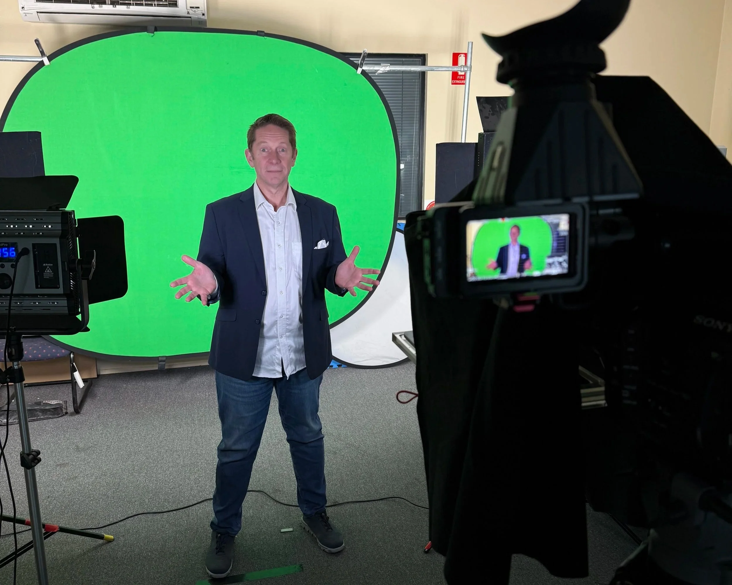 A man in a dark blazer and white shirt stands in front of a green screen, with video recording equipment around him, speaking to the camera in an indoor setting.