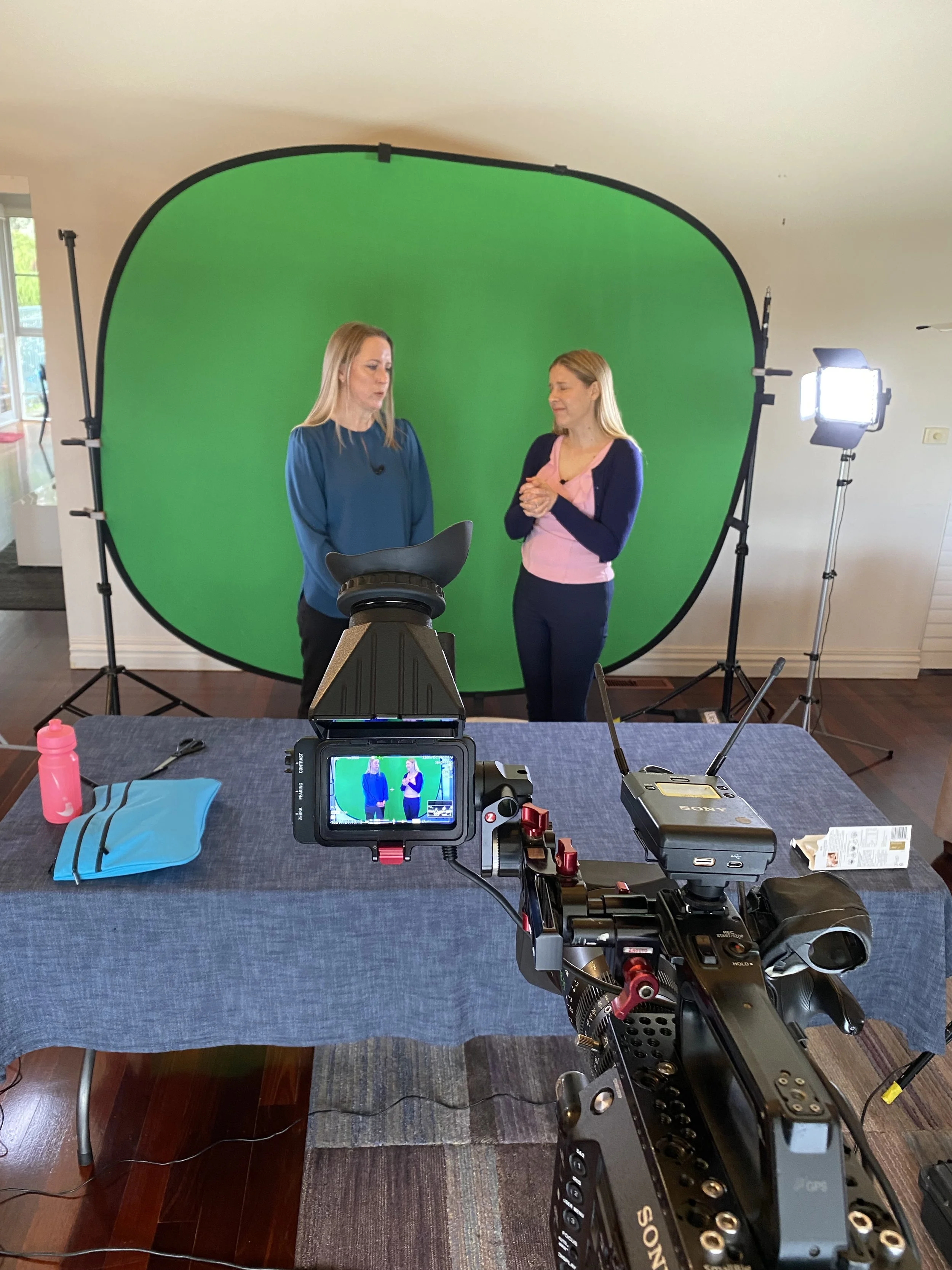 A camera filming two women standing in front of a green screen with lighting equipment set up in a room.